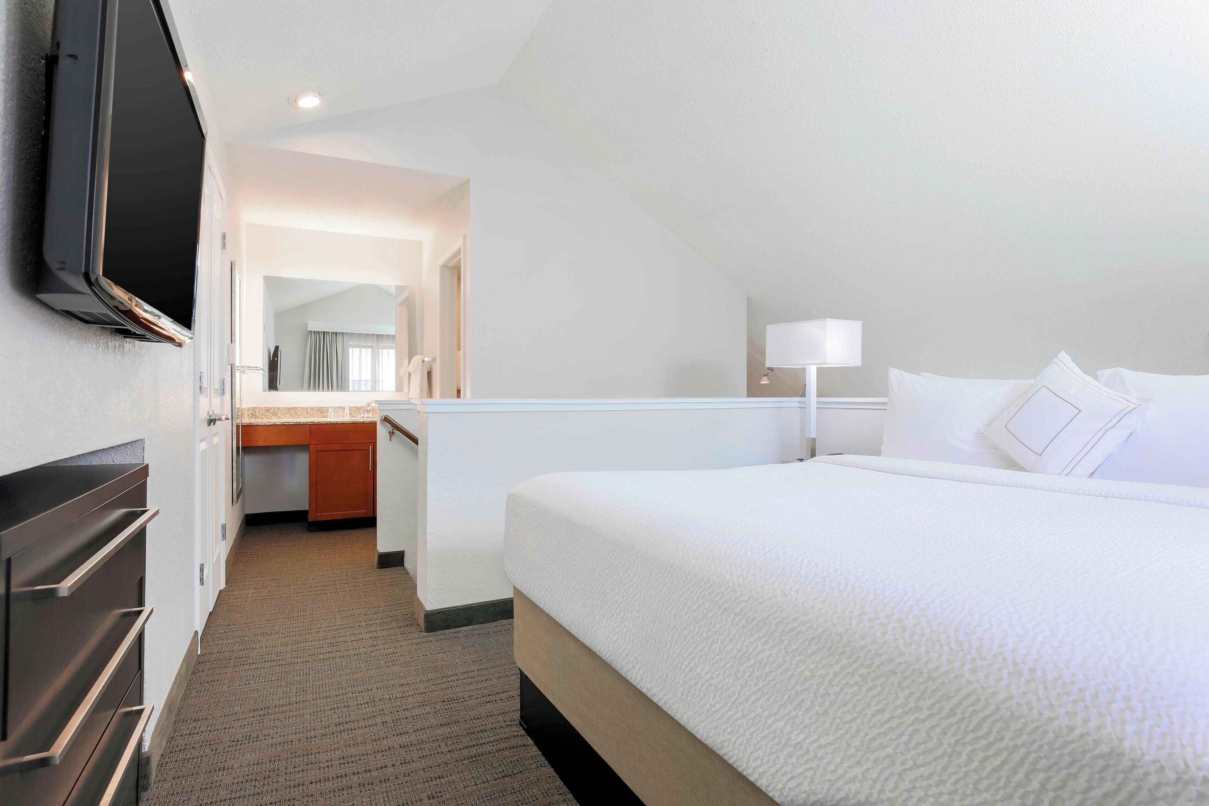 Habitación de hotel con cama, televisor montado en la pared, escritorio con lámpara y baño visible a través de una puerta abierta.