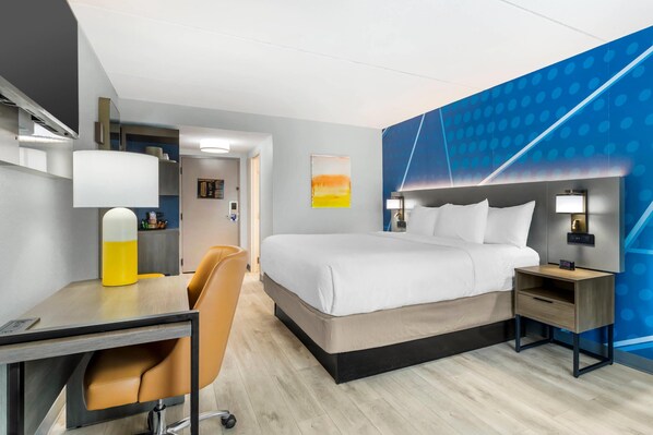 A hotel room with a bed, bedside table, lamp, desk, chair, and a wall with a geometric pattern.