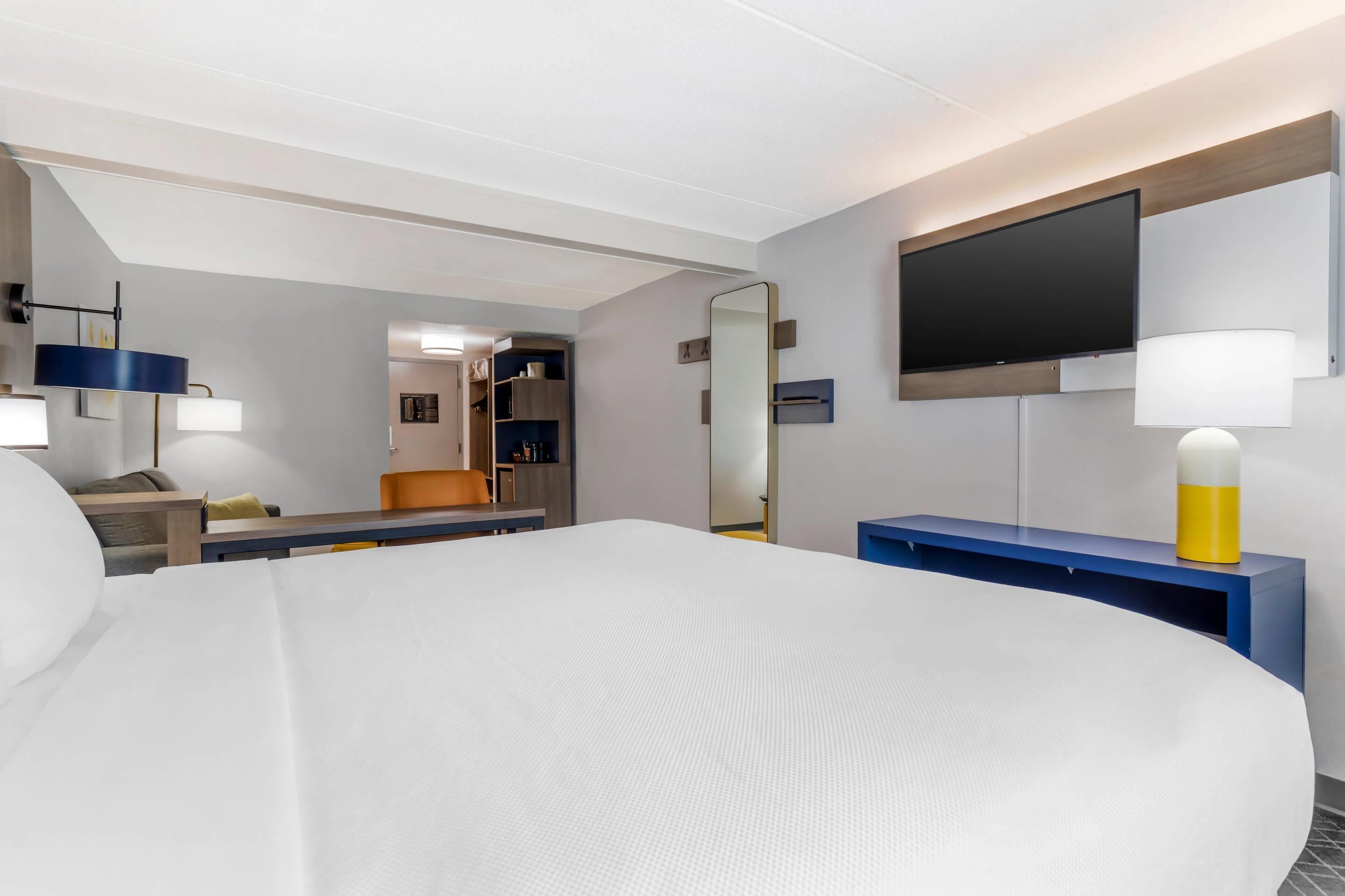 Et moderne hotelværelse med en stor seng, et fladskærms-tv på væggen, et skrivebord med en lampe og et spejl.