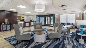 A modern hotel lobby with grey armchairs, a round coffee table, and a blue patterned rug. There is a reception desk in the background and a blue sofa with a lamp.