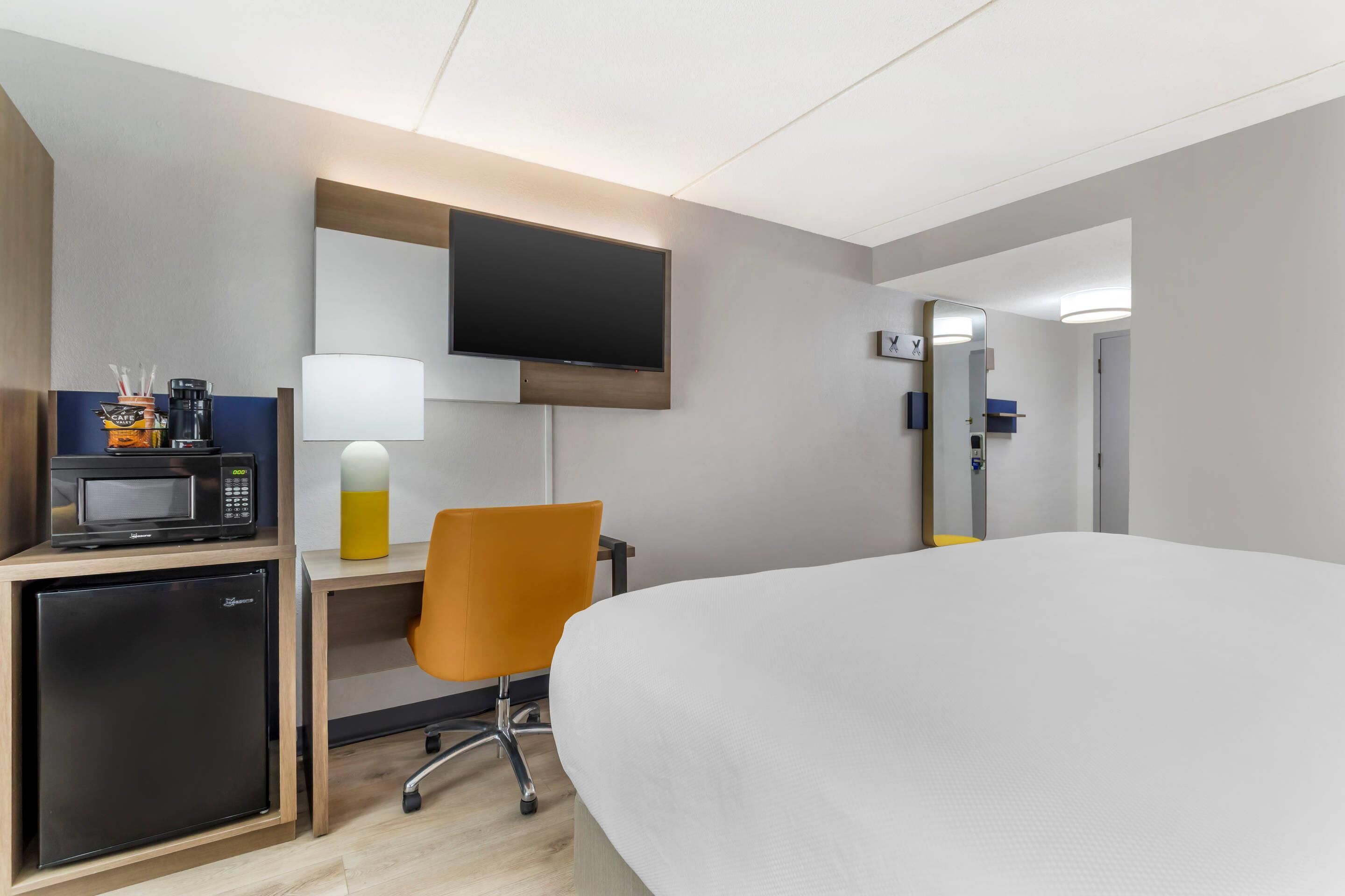 Ein Hotelzimmer mit einem Bett, einem Schreibtisch mit Mikrowelle, einem Sessel, einem an der Wand montierten Fernseher und einem kleinen Kühlschrank.