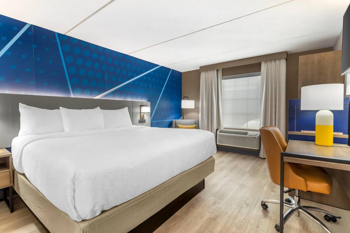 A hotel room with a large bed, a desk, a chair, a desk lamp, a window with curtains, and a wall with a blue pattern.