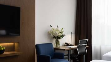 A hotel room with a flat-screen TV mounted on the wall, a desk with a telephone and a vase of flowers, a blue armchair, and a bed with white bedding.