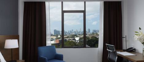 A hotel room with a bed, a desk, a chair, a lamp, and a view of a cityscape through large windows.