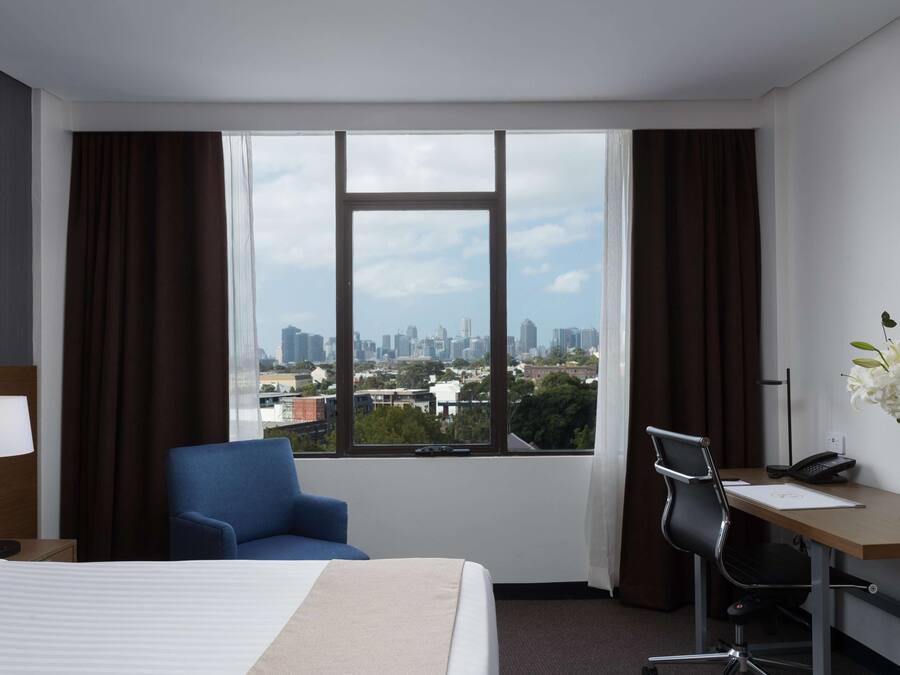 Habitación de hotel con cama, escritorio, silla, lámpara y vista de un paisaje urbano a través de amplias ventanas.