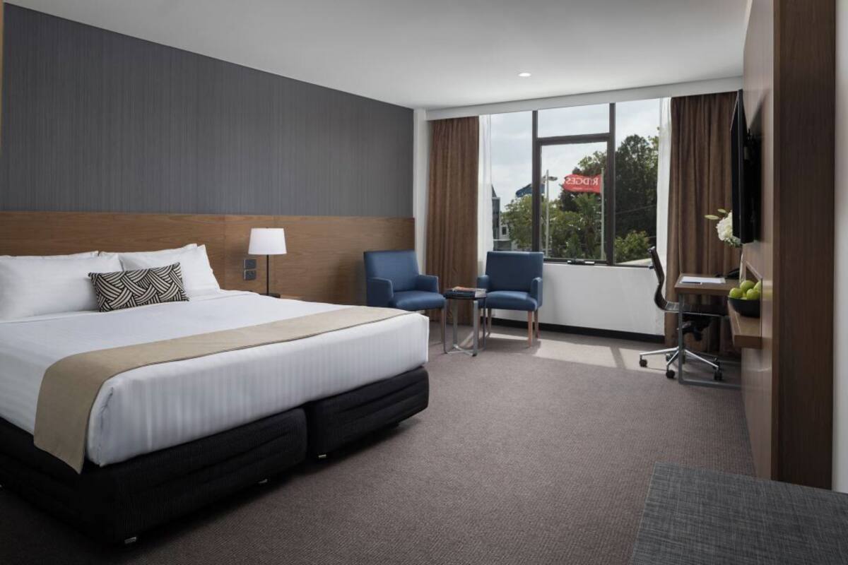 Et hotelværelse med en stor seng, to lænestole, et skrivebord med en bærbar computer og et fjernsyn.