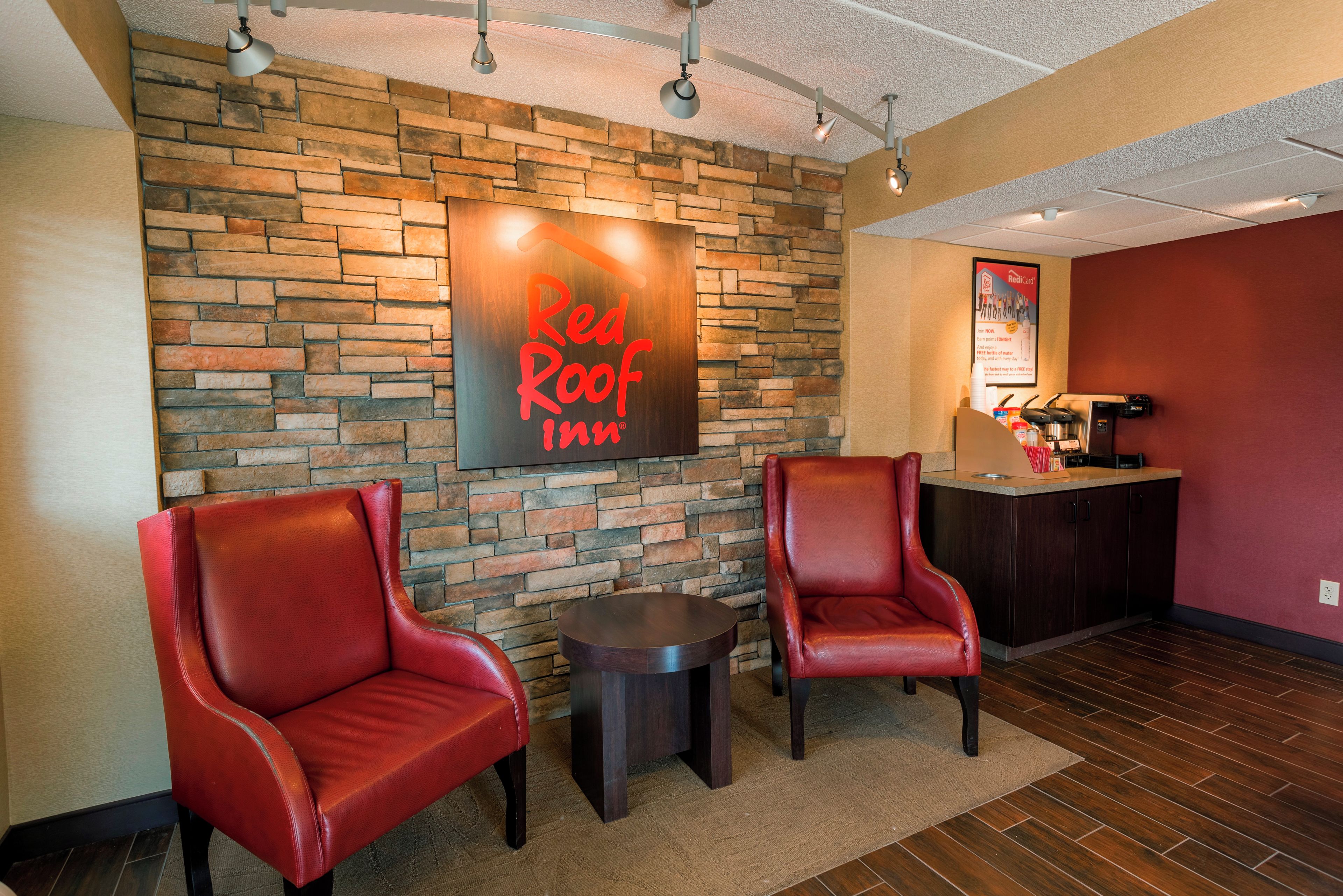 A Red Roof Inn lobby with a stone wall, two leather chairs, a small table, and a reception desk with amenities.