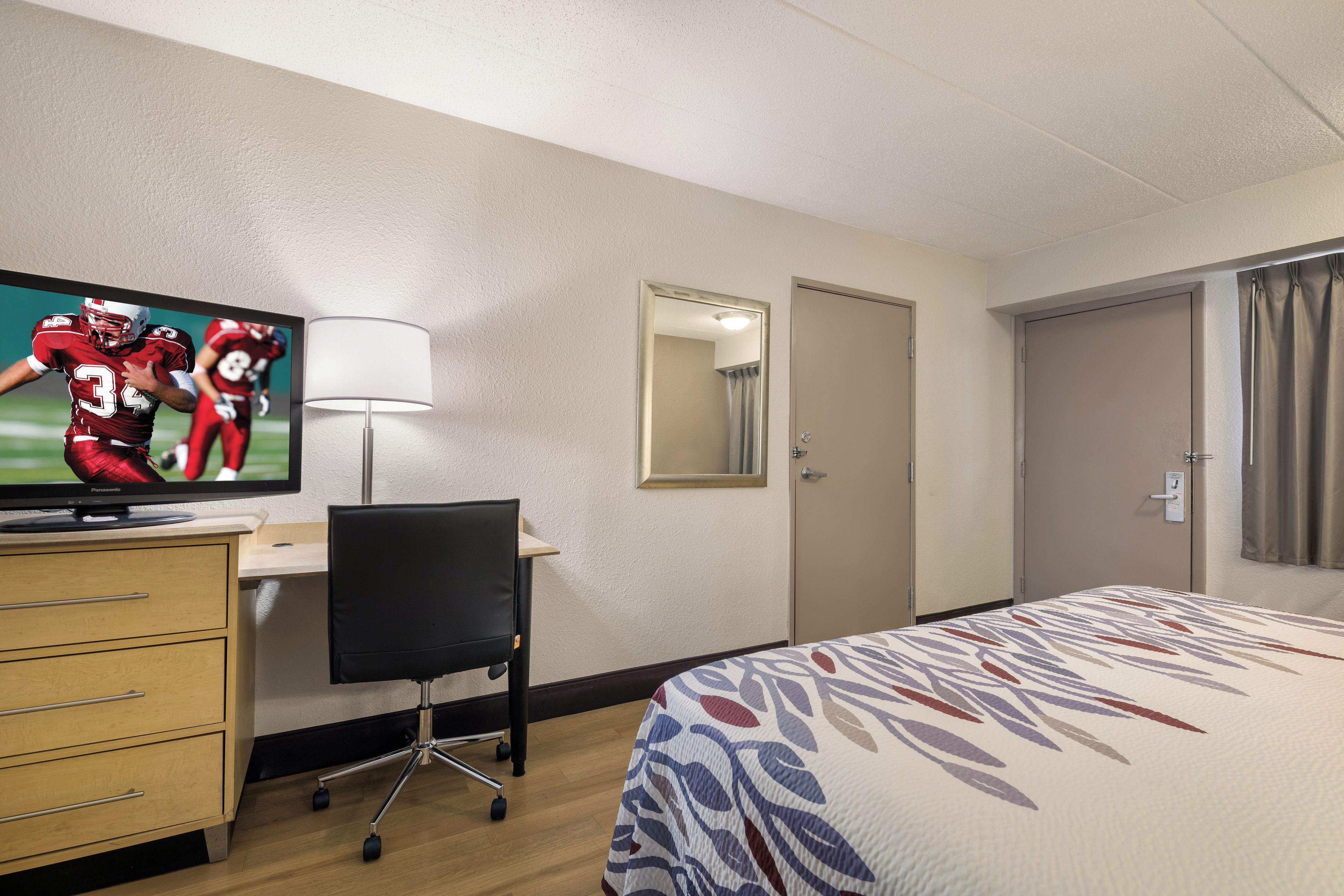 A hotel room with a bed, a desk, a television, a chair, a lamp, a door, and a mirror.