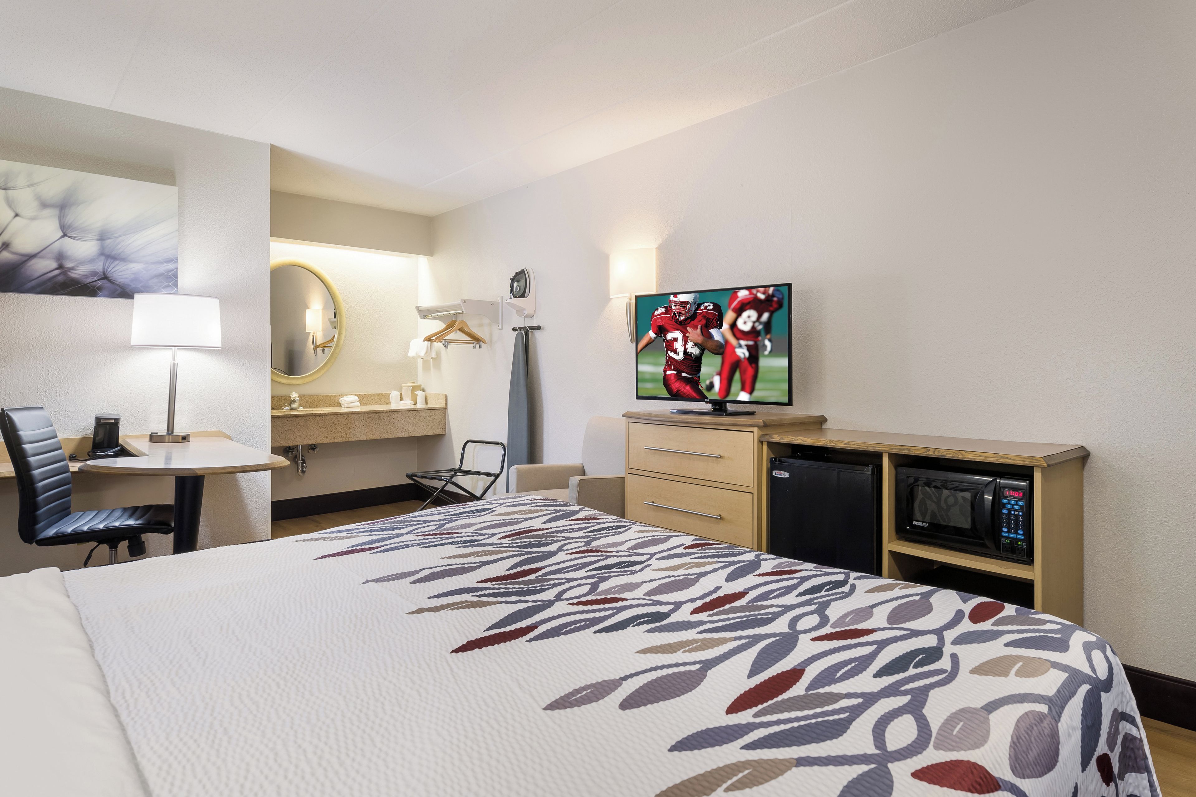 A hotel room with a bed, a desk with a chair, a television, a mirror, and a microwave.
