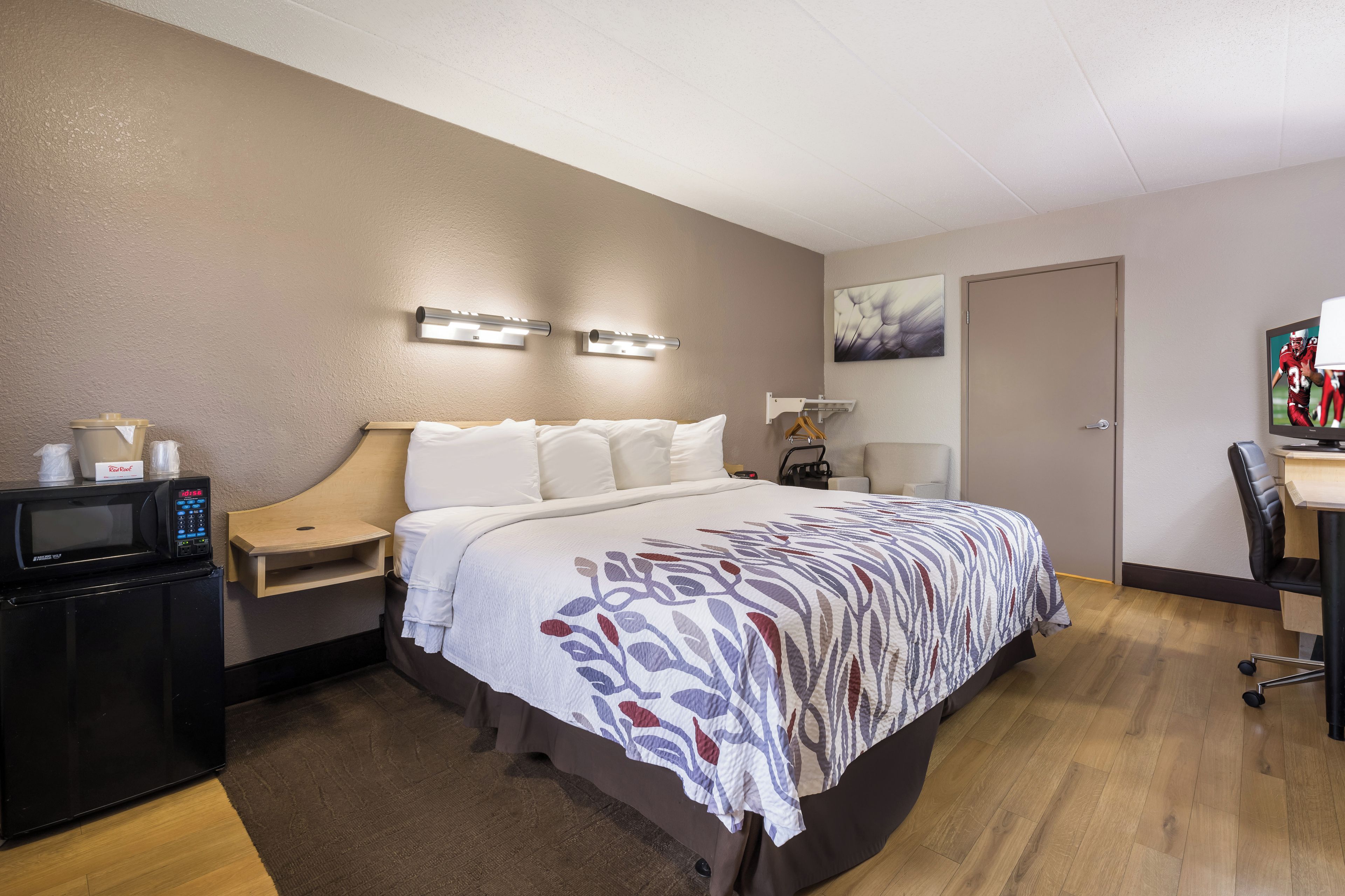 A hotel room with a bed, bedside table, a microwave, a desk with a chair, and a door.