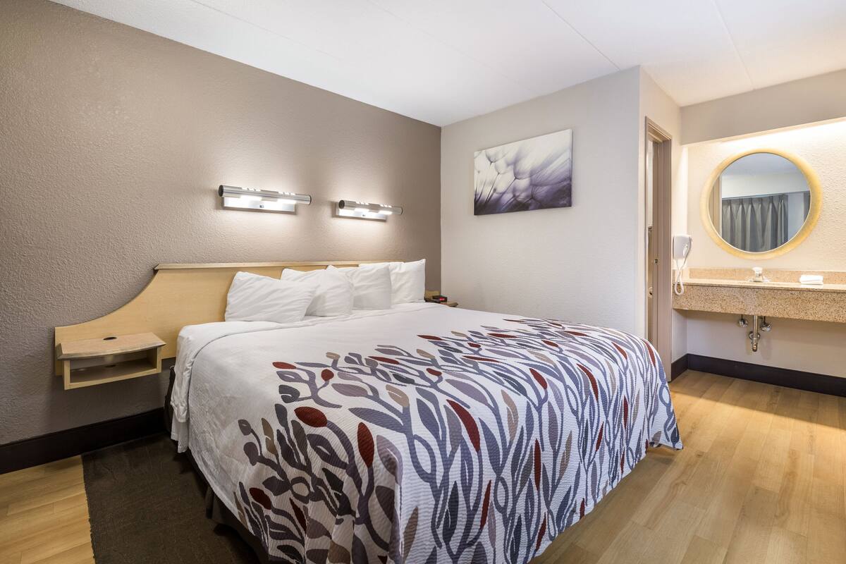 Ein Hotelzimmer mit einem großen Bett, Nachttischen, einer Wandleuchte, einem Gemälde und einem Badezimmer mit Waschbecken und Spiegel.