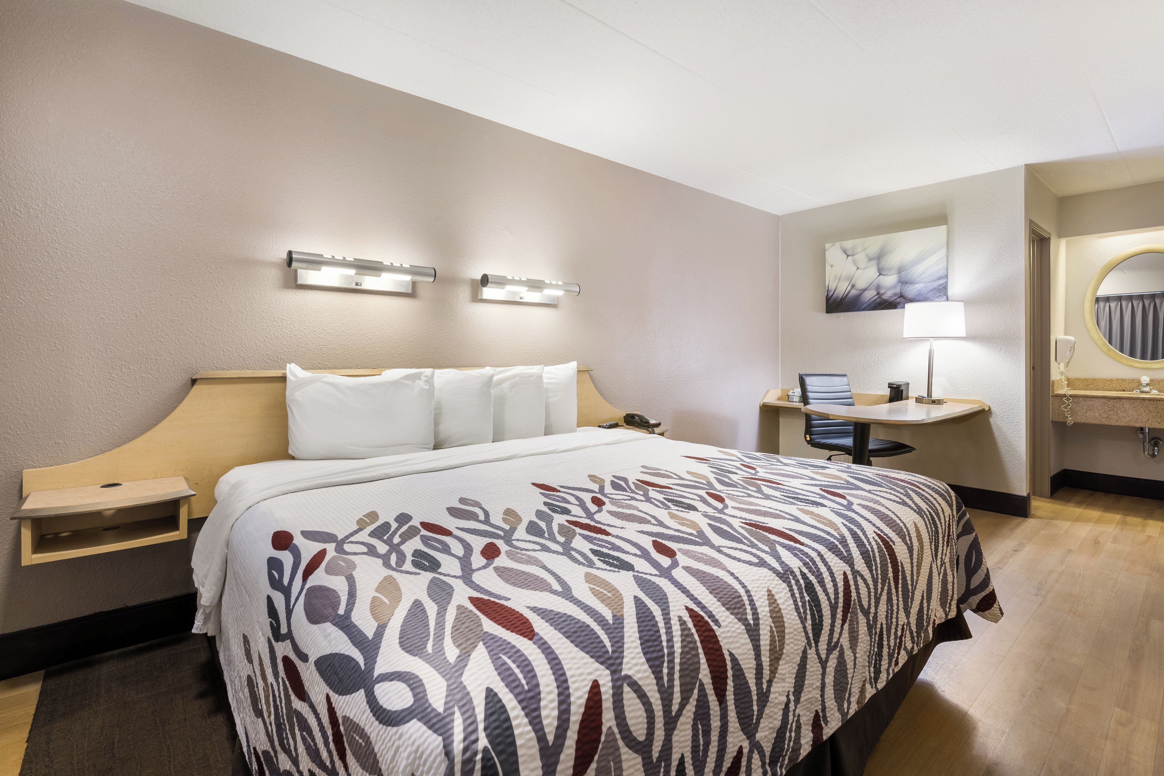 Une chambre d’hôtel comprenant un lit, une table de chevet, un bureau avec une chaise, une salle de bain avec un miroir et un tableau accroché au mur.