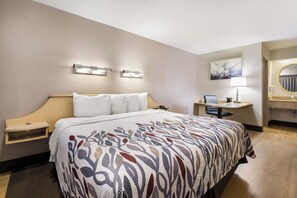 A hotel room with a bed, bedside table, a desk with a chair, a bathroom with a mirror, and a painting on the wall. - Red Roof Inn Rockford East – Casino District (Rockford)