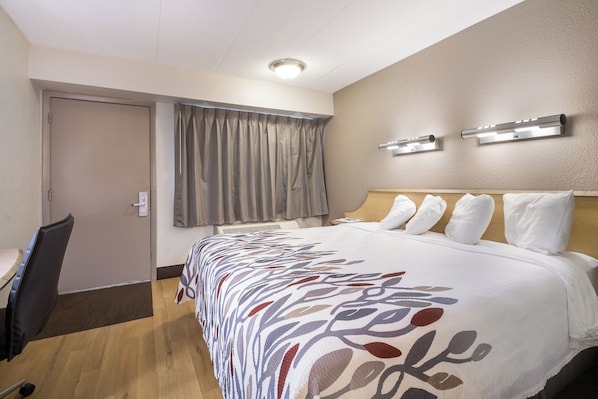 A hotel room with a bed, a desk, a chair, a window with curtains, and an air conditioning unit.