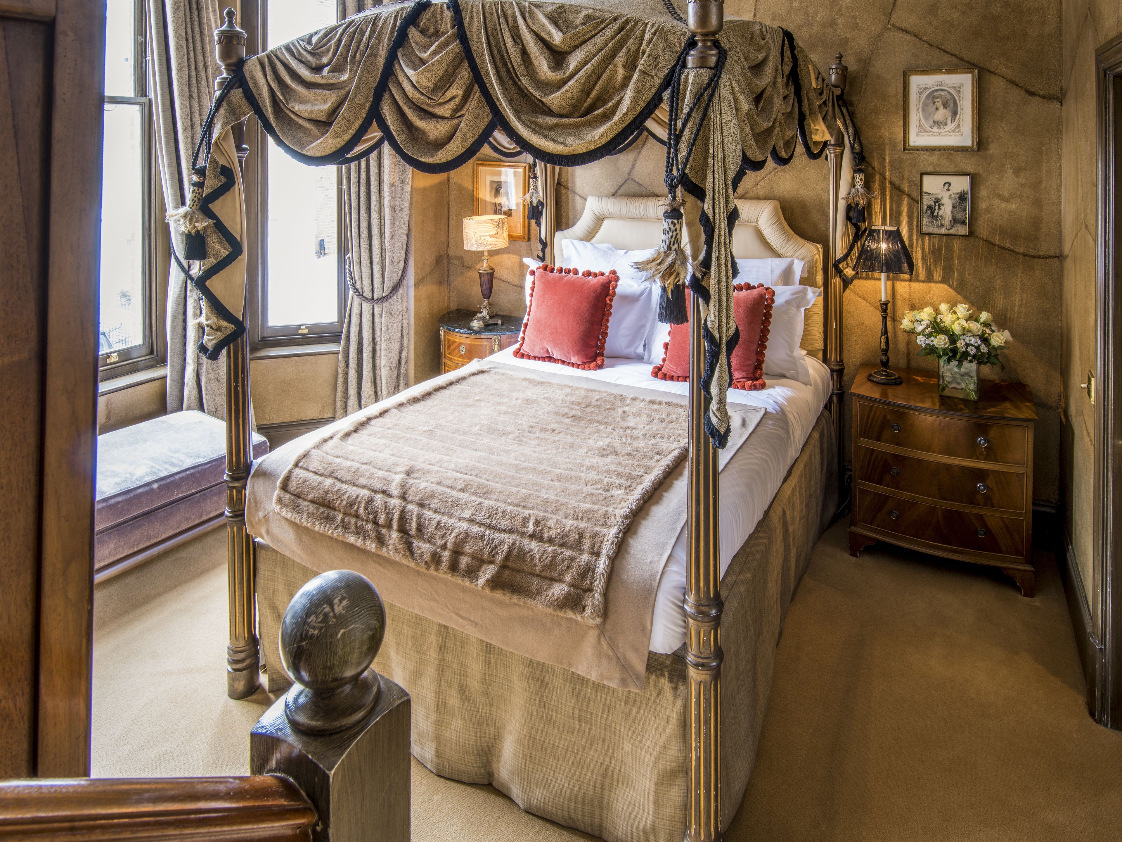 A four-poster canopy bed with a wooden headboard, a bedside table with a lamp, and a nightstand with a vase of flowers.