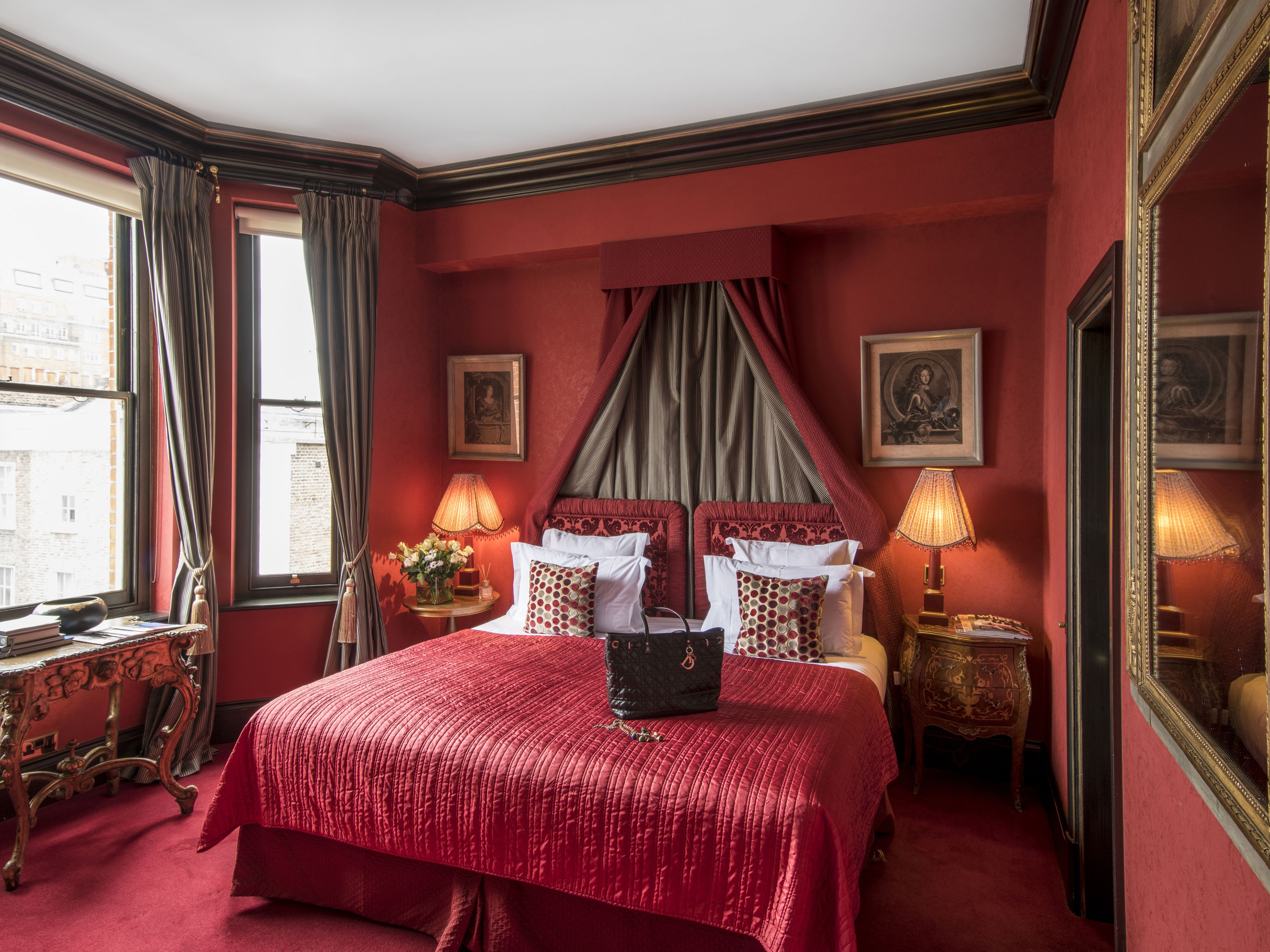 A red-themed bedroom with a large bed, two bedside tables with lamps, a mirror, and a window with curtains.