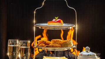 A three-tiered tea service set with sandwiches and pastries, surrounded by tea cups and a teapot, with a fireplace in the background.