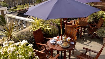 A terrace with wooden furniture, a table set with a teapot, cups, and a plate of food, under a large umbrella.