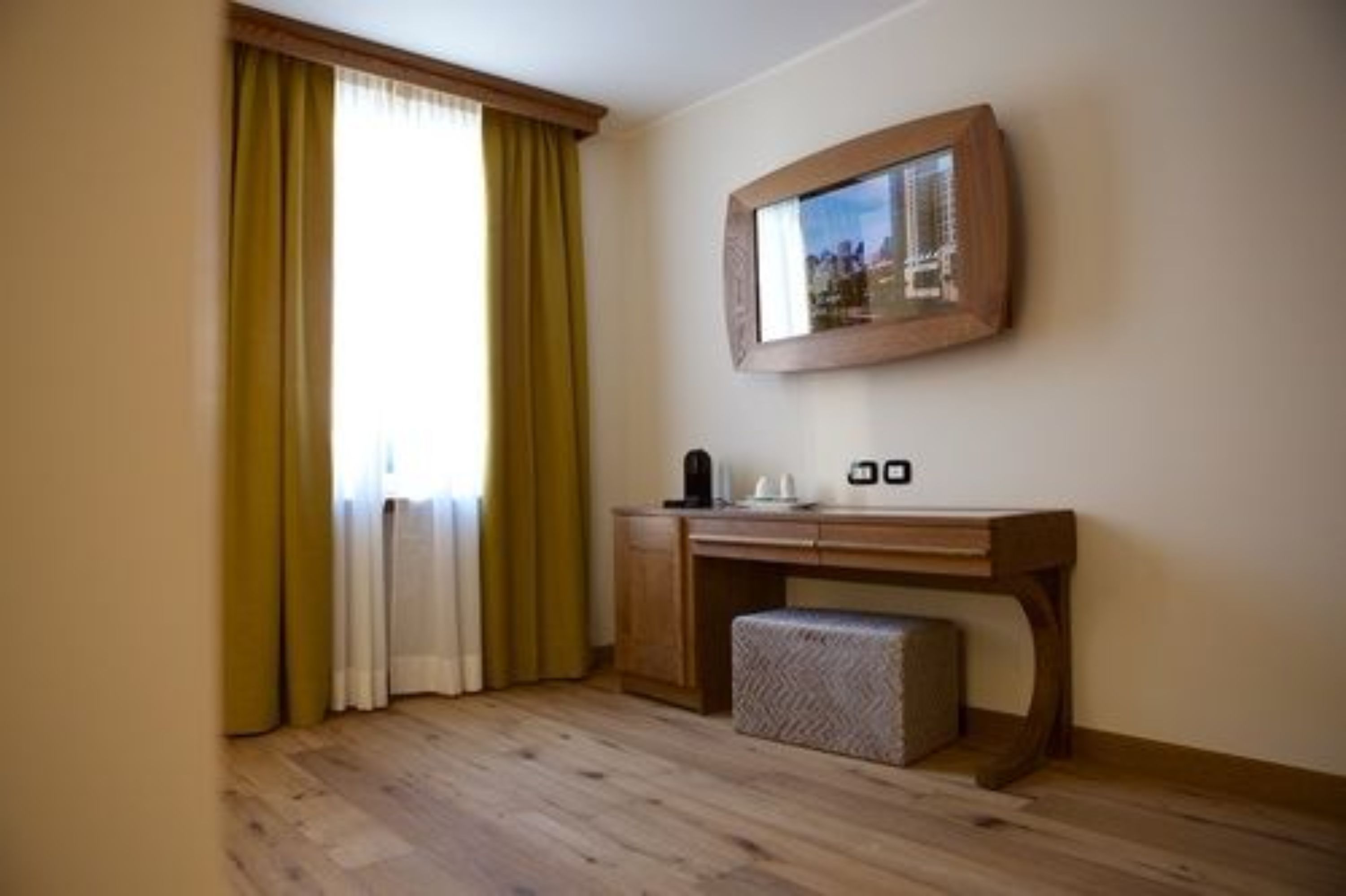 Une pièce avec un bureau en bois, un petit banc, un tableau encadré au mur et une fenêtre avec des rideaux.