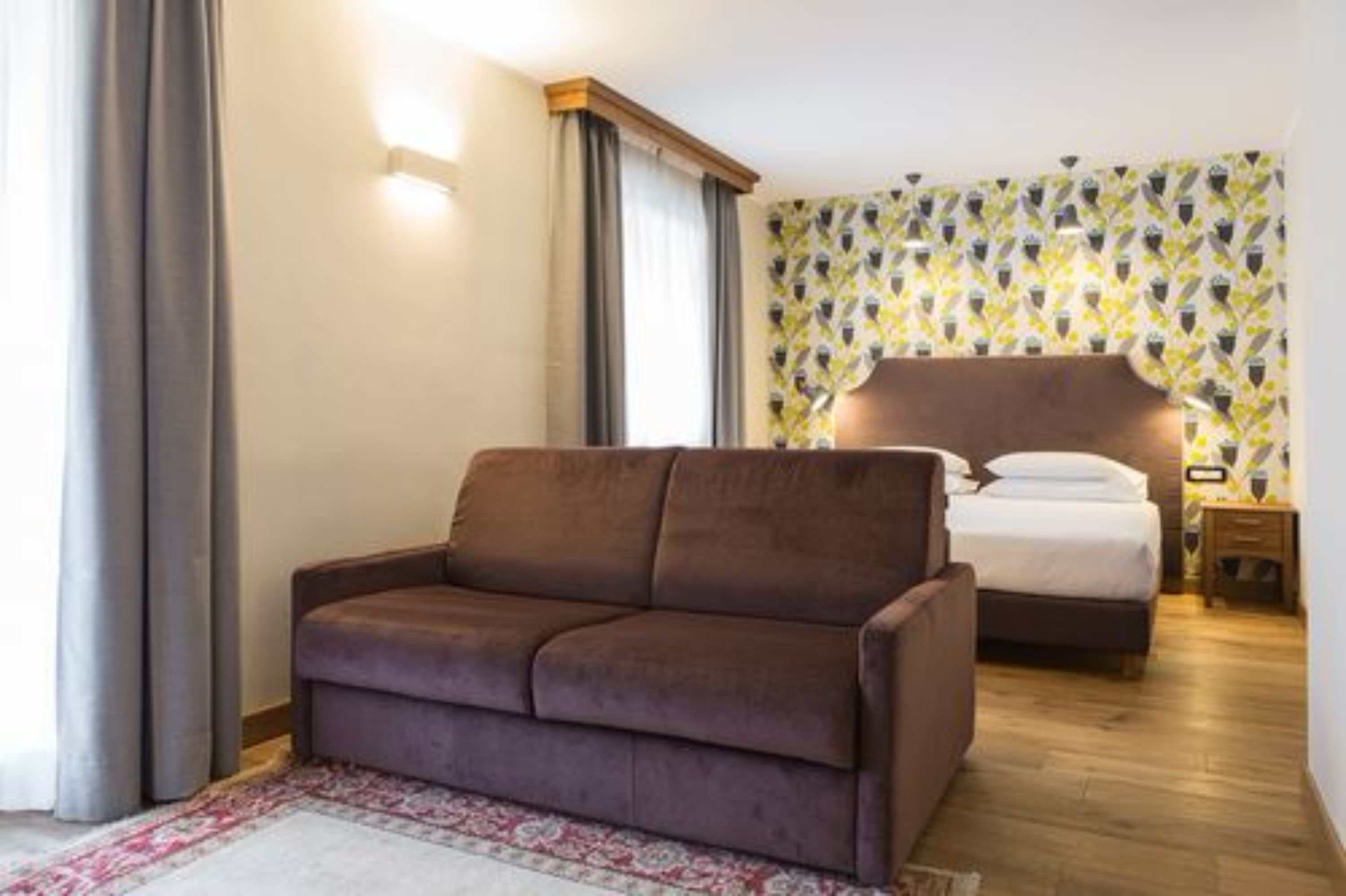 A bedroom with a brown sofa, a bed with a wooden headboard, a nightstand, and a patterned wall.