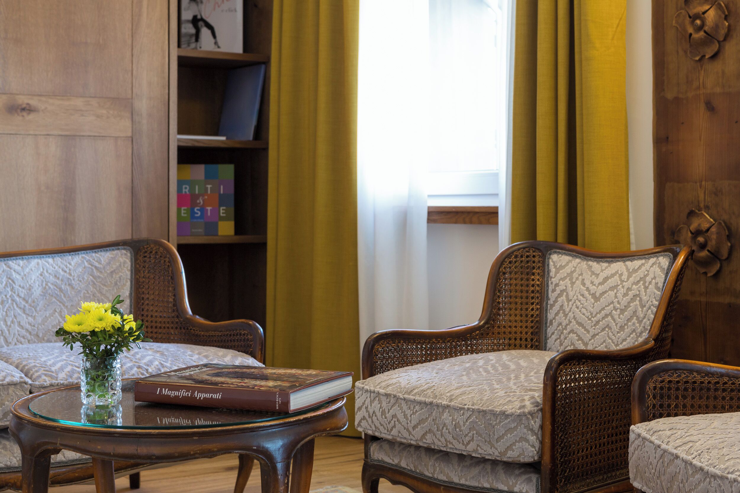 A room with a wooden bookshelf, a round table with a book and a vase, and two upholstered chairs.