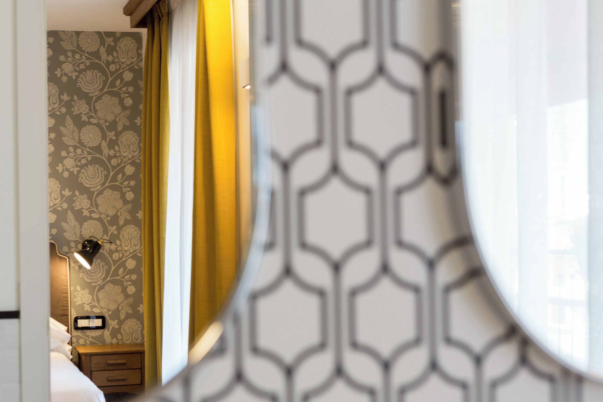 A room with a patterned wallpaper, a bedside lamp, a nightstand, and a mirror with a geometric design.