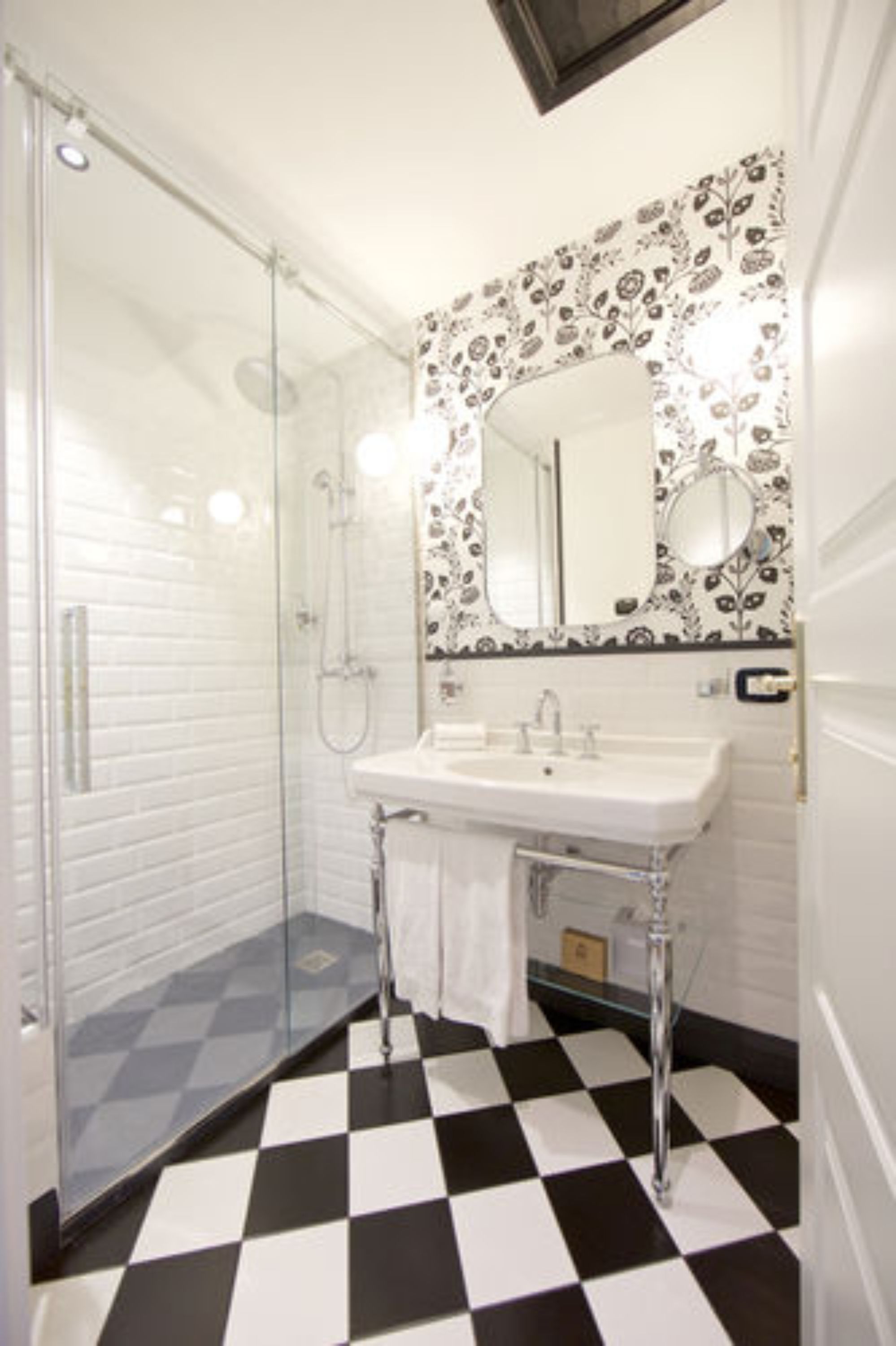 Un baño con piso a cuadros, cabina de ducha de vidrio, lavamanos blanco y espejo con papel tapiz floral.