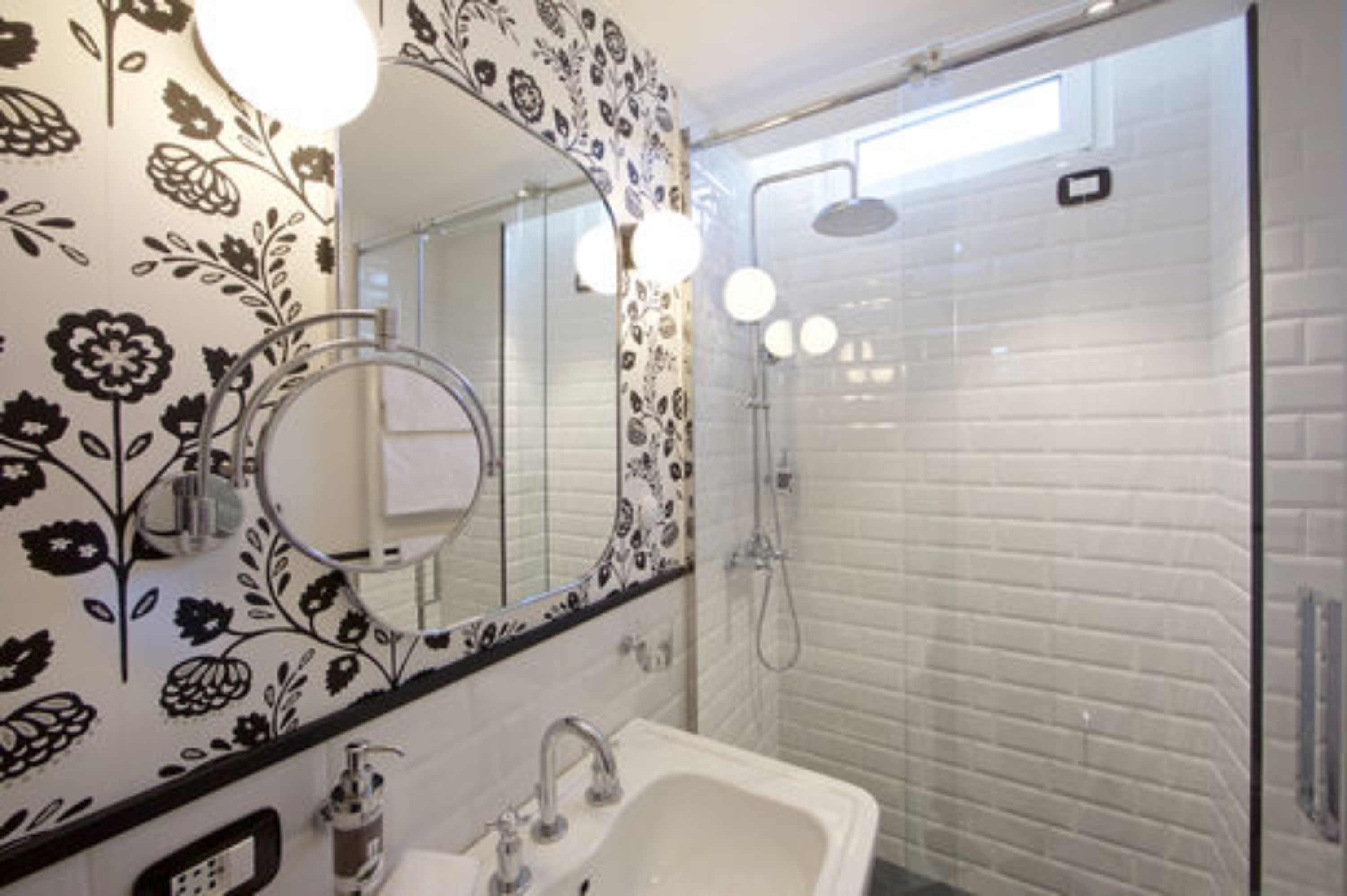 A bathroom with a floral-patterned wall, a round mirror, a sink, and a shower area.