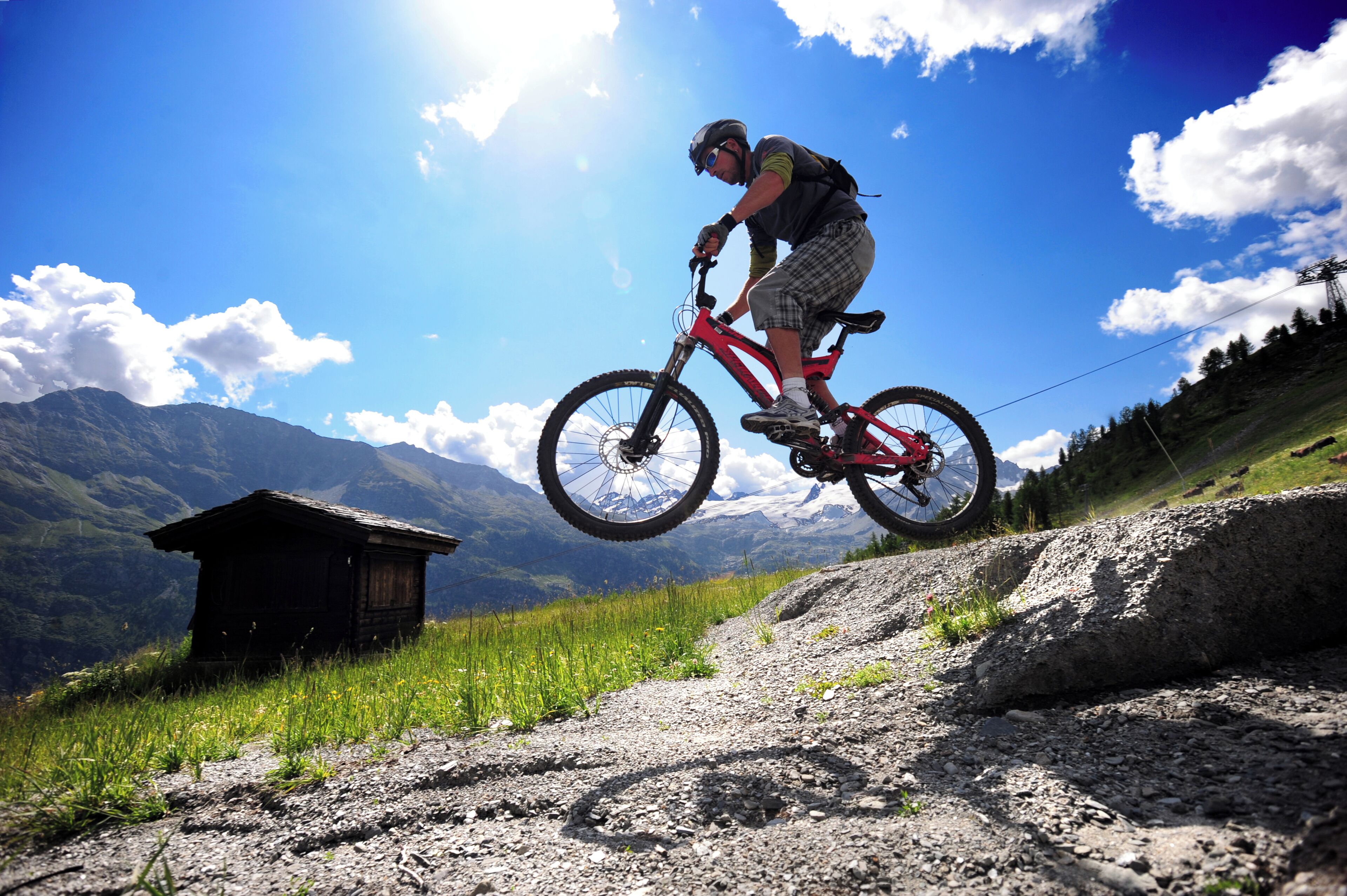 Un ciclista da mountain bike esegue un salto sopra un terreno roccioso, con una struttura in legno sullo sfondo e un paesaggio montuoso.