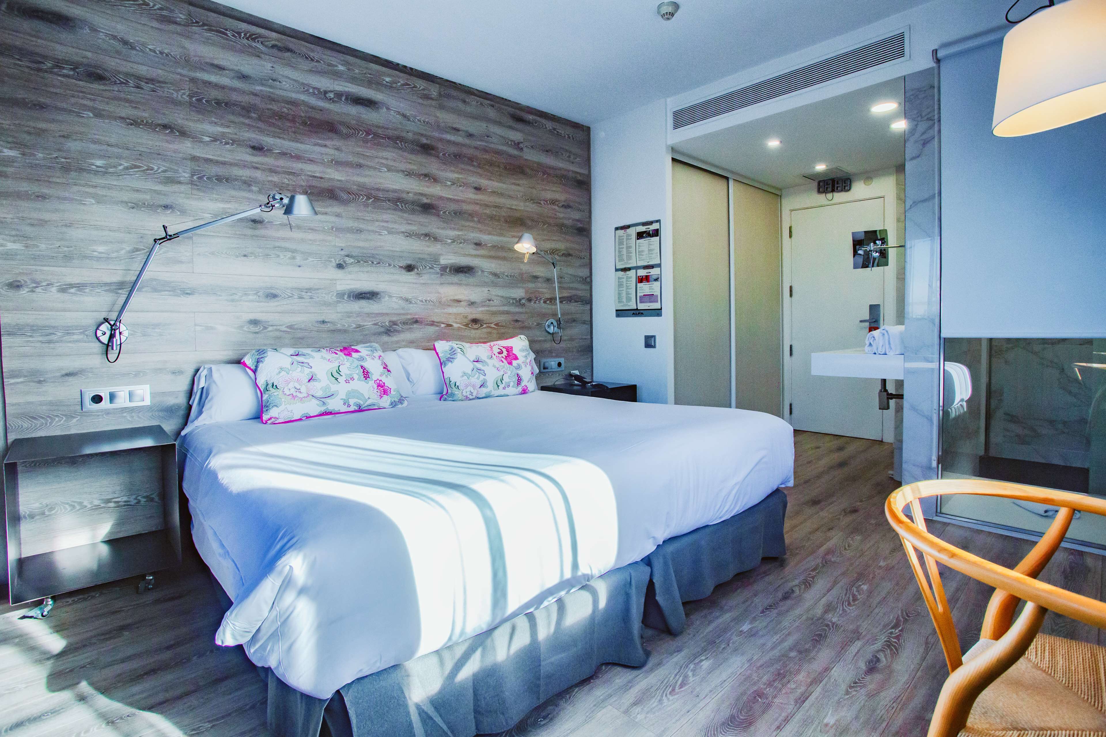 A modern hotel room with a wooden headboard, a large bed with white bedding, a bedside table, a chair, and a bathroom visible through a glass door.