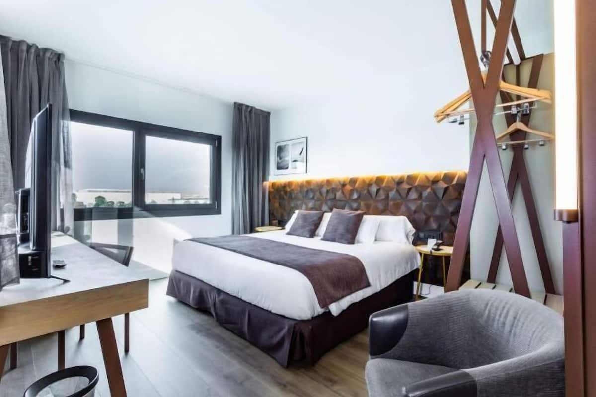 Ein modernes Hotelzimmer mit einem großen Bett, einem Schreibtisch, einem Sessel und Blick durch ein Fenster.