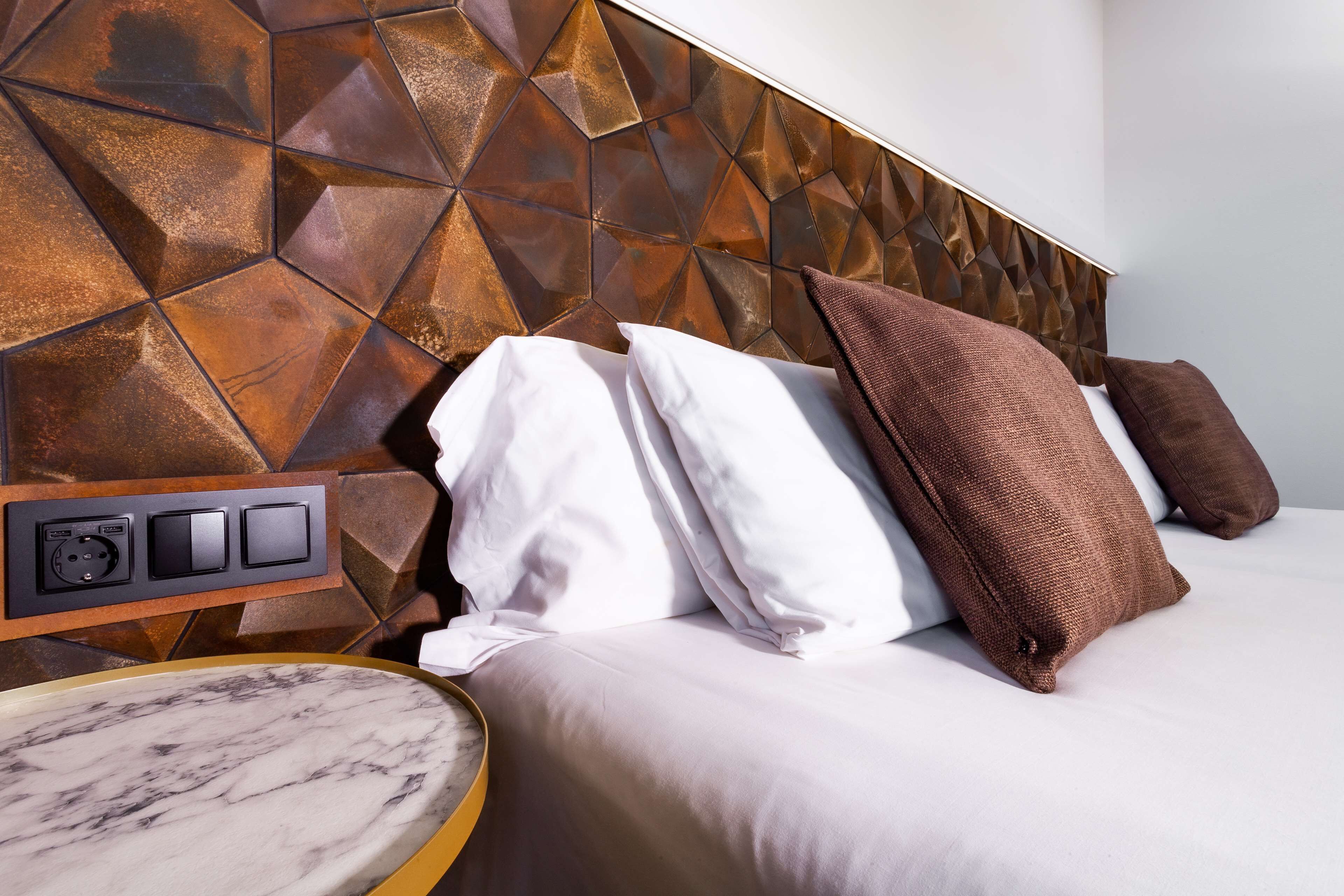 A modern hotel room with a geometric-patterned headboard, a marble bedside table, and a neatly made bed with white and brown pillows.