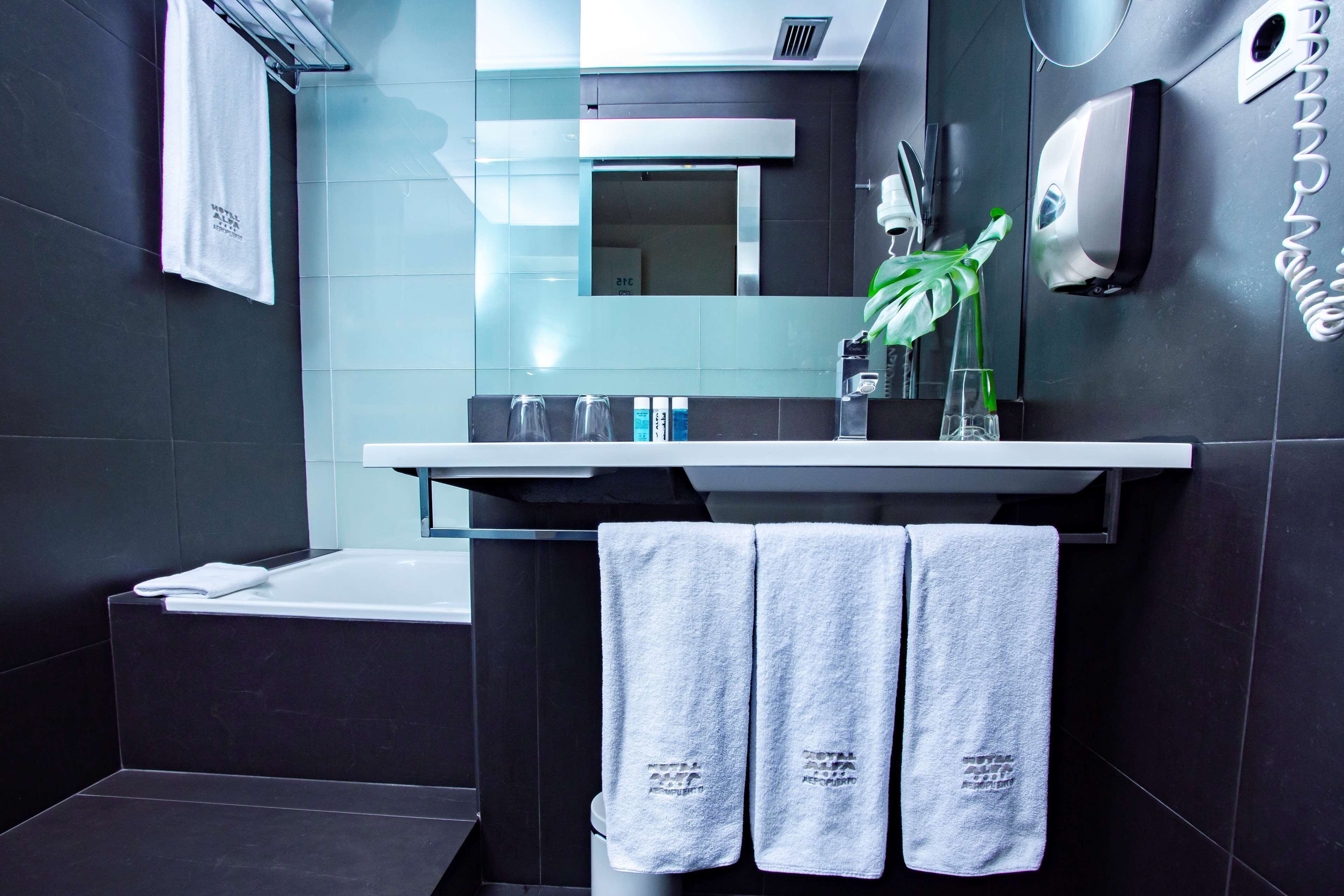 A modern bathroom with a bathtub, a towel rack with three towels, a hand dryer, and a mirror.