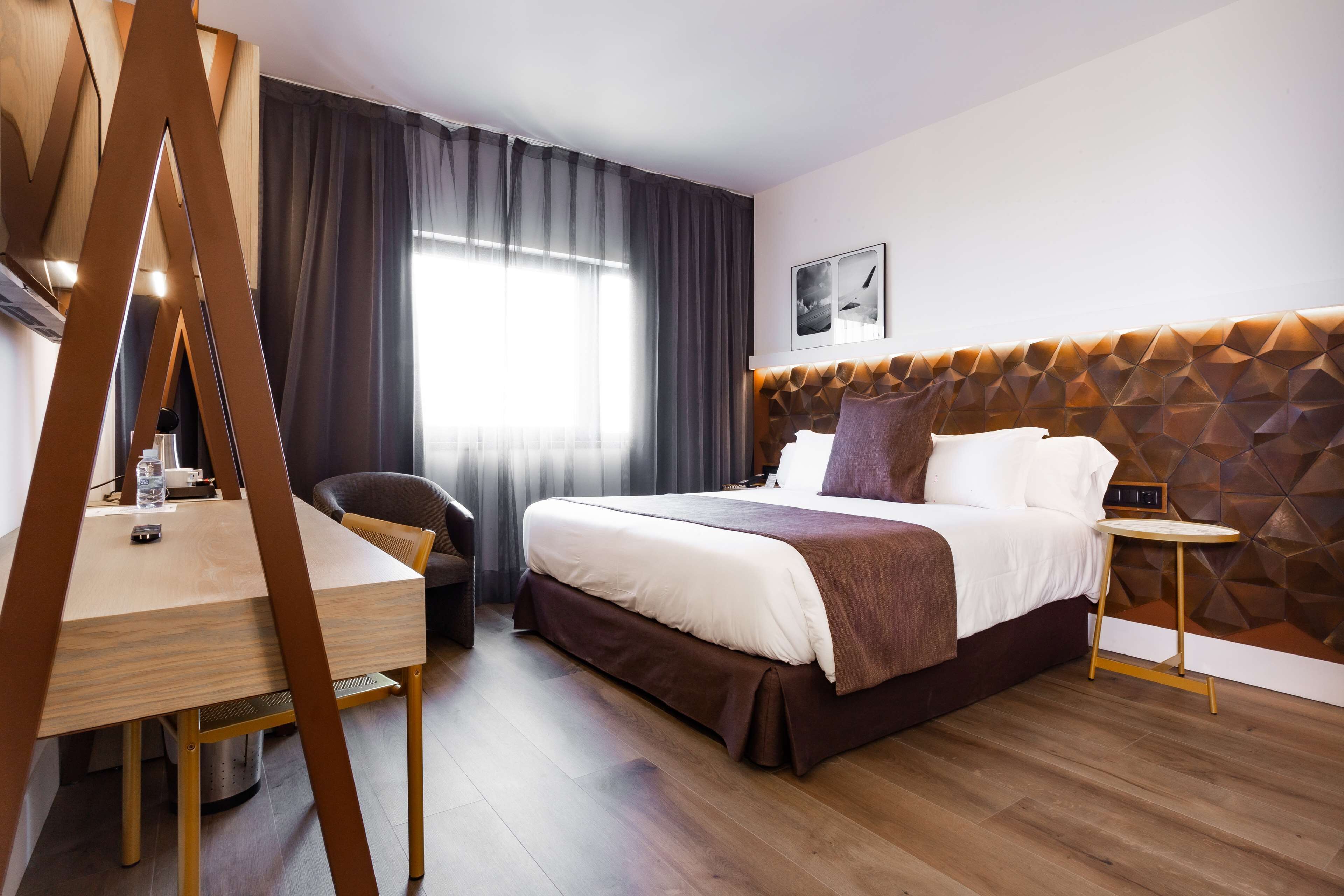 Habitación de hotel con cama, escritorio, silla, mesita y ventana con cortinas.