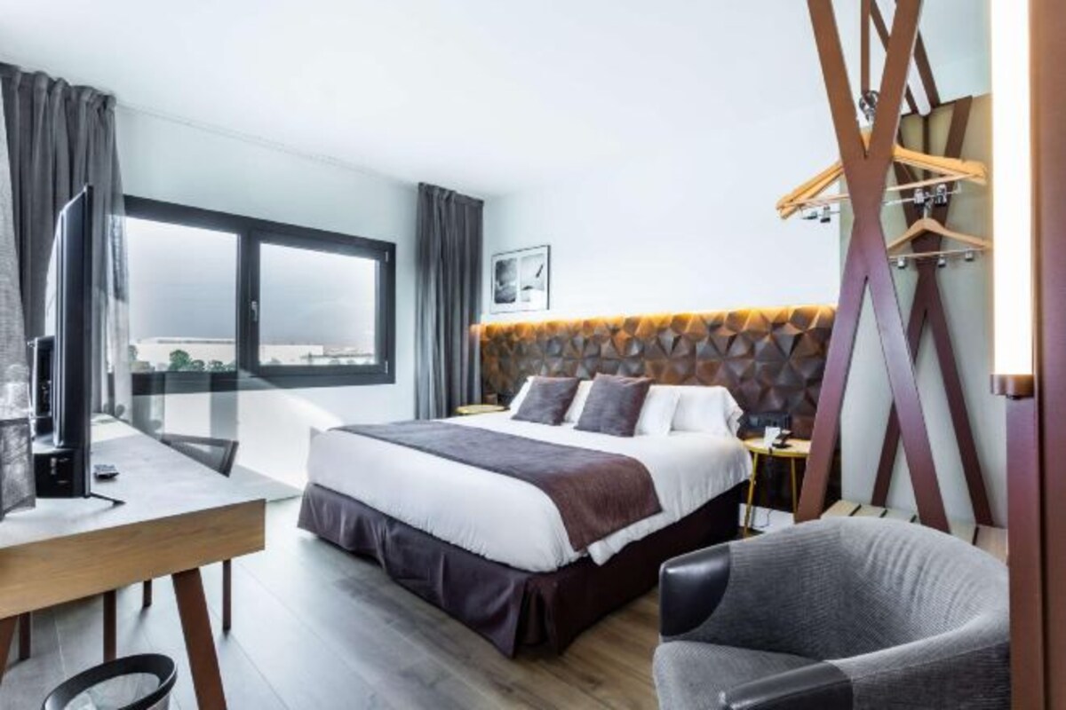 A modern hotel room with a large bed, a desk, a chair, and a view through a window.