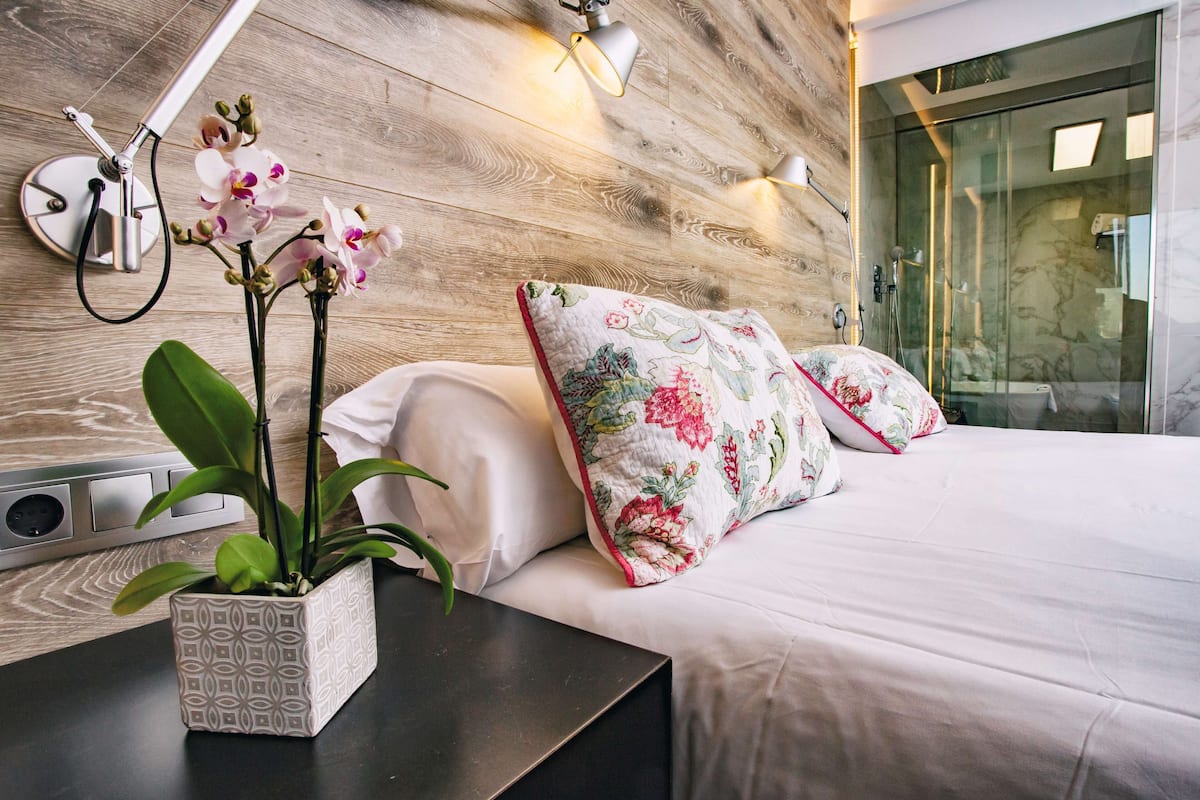 Ein modernes Schlafzimmer mit einem hölzernen Kopfteil, einem Nachttisch mit einer Zierpflanze und einem Bett mit blumigen Kissen.