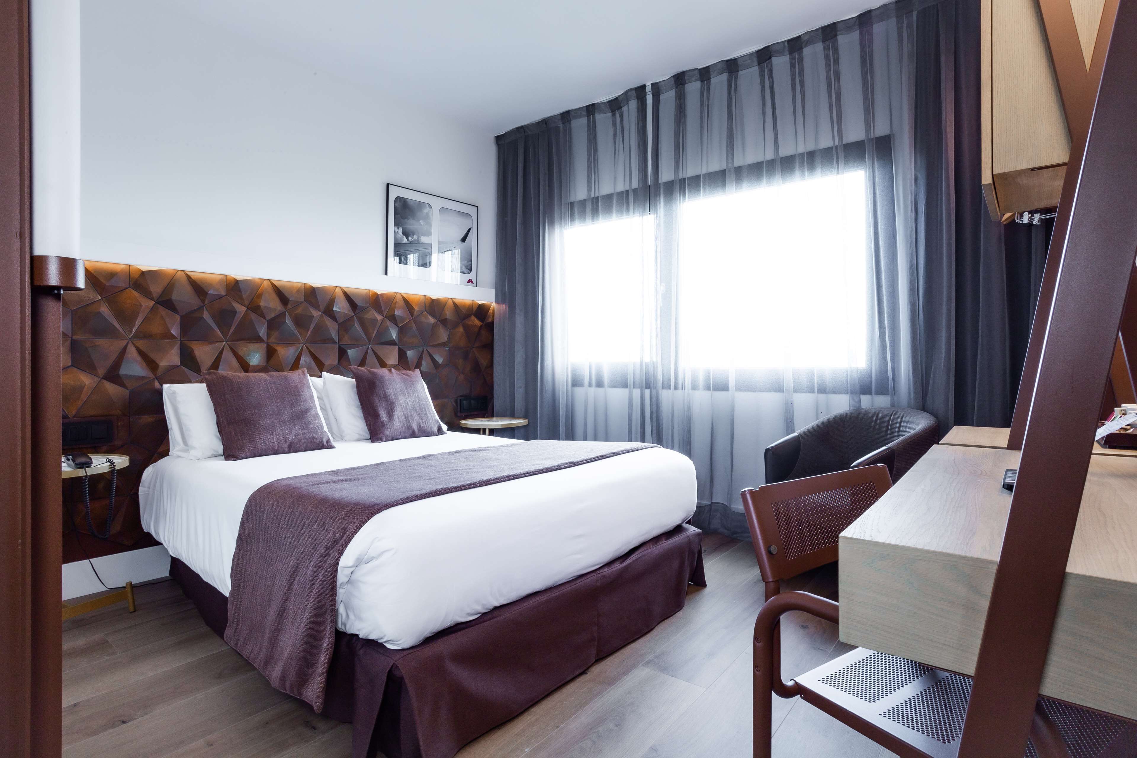A modern hotel room with a geometric-patterned headboard, a large bed with white and brown bedding, a desk with a chair, and a large window with sheer curtains.