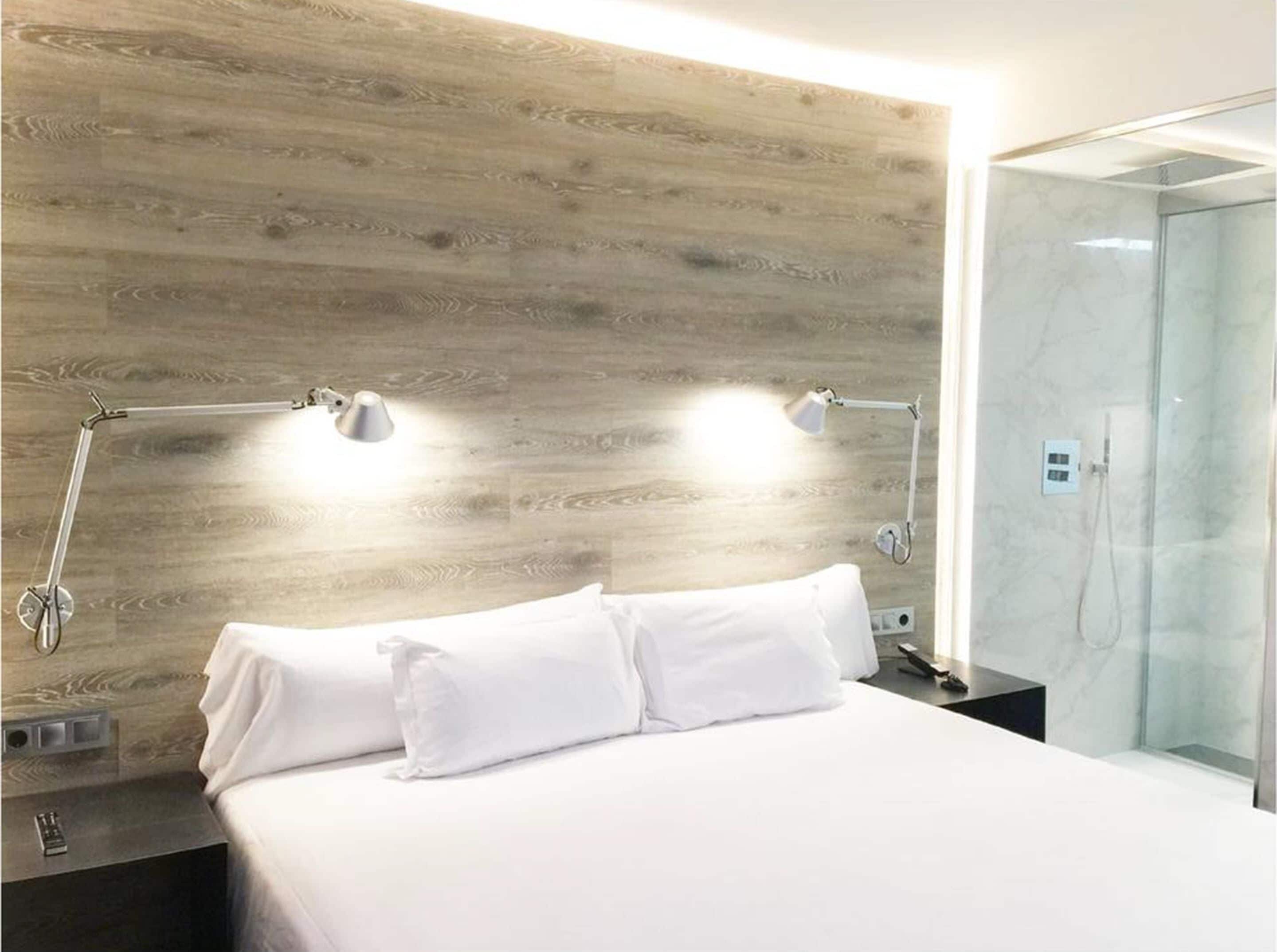 Een moderne hotelkamer met een houten hoofdbord, een bed met witte lakens, twee bedlampjes, een glazen douchecabine en een klein nachtkastje.