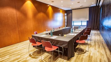 Uma sala de conferências com uma mesa comprida, cadeiras vermelhas e uma tela de projetor.