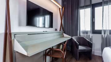 Habitación de hotel moderna con una televisión de pantalla plana montada en la pared, un escritorio de madera, un sillón oscuro y un gran ventanal con cortinas.