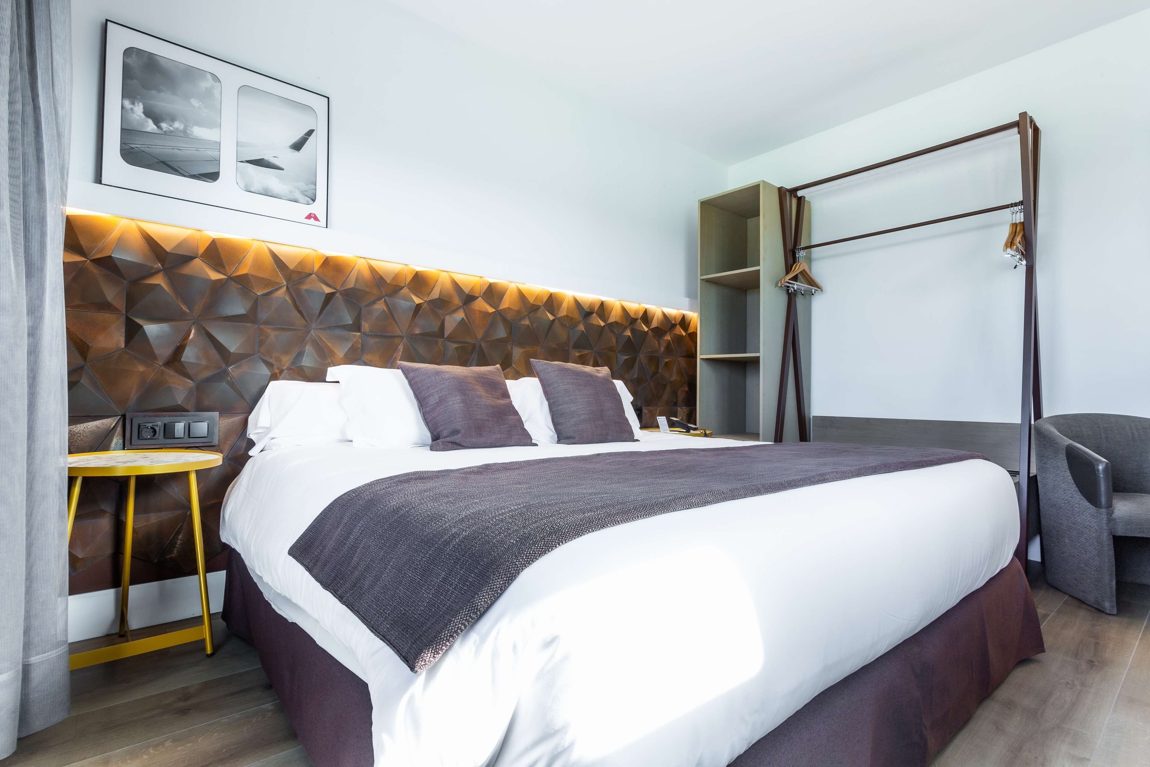 Quarto de hotel moderno com cabeceira estampada em padrão geométrico, uma cama grande com roupa de cama branca e marrom, uma pequena mesa de apoio, uma cadeira e um guarda-roupa com espelho.