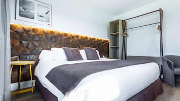 Quarto de hotel moderno com cabeceira estampada em padrão geométrico, uma cama grande com roupa de cama branca e marrom, uma pequena mesa de apoio, uma cadeira e um guarda-roupa com espelho.
