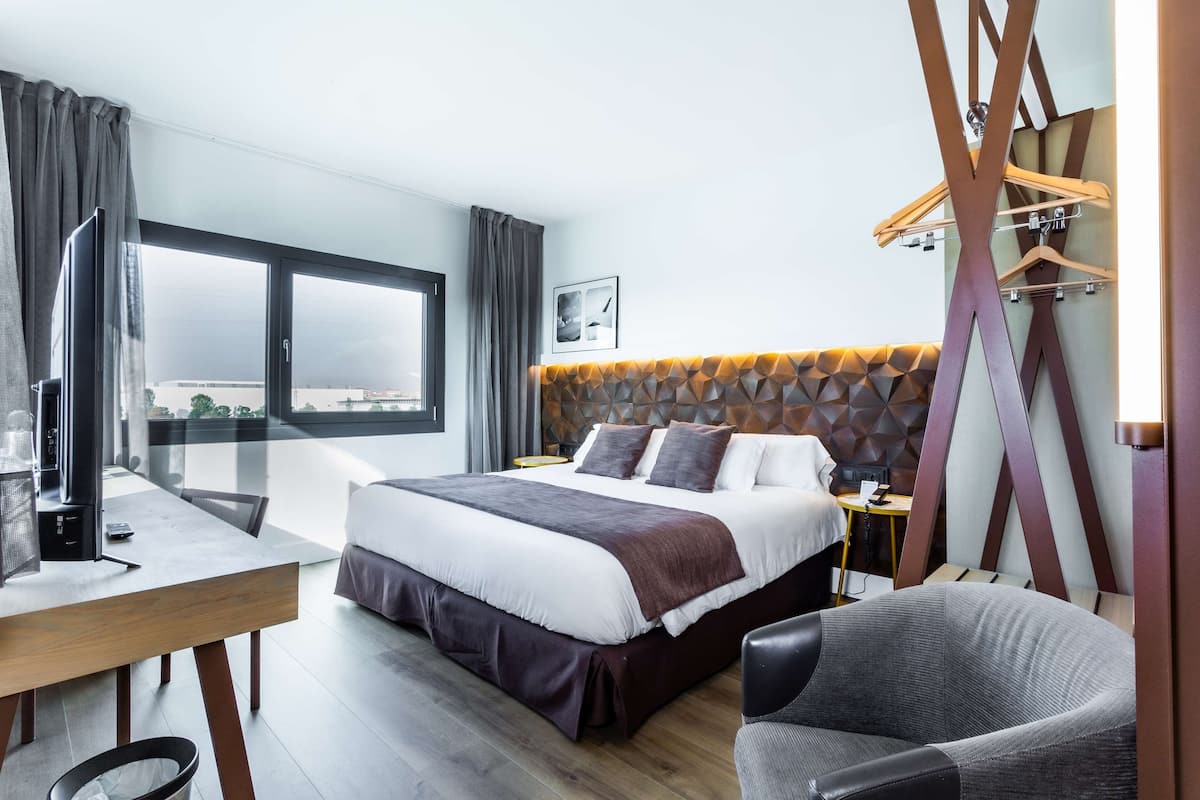 Ein modernes Hotelzimmer mit einem großen Bett, einem Schreibtisch und einem Sessel. Ein Fenster bietet Blick auf die Stadt.