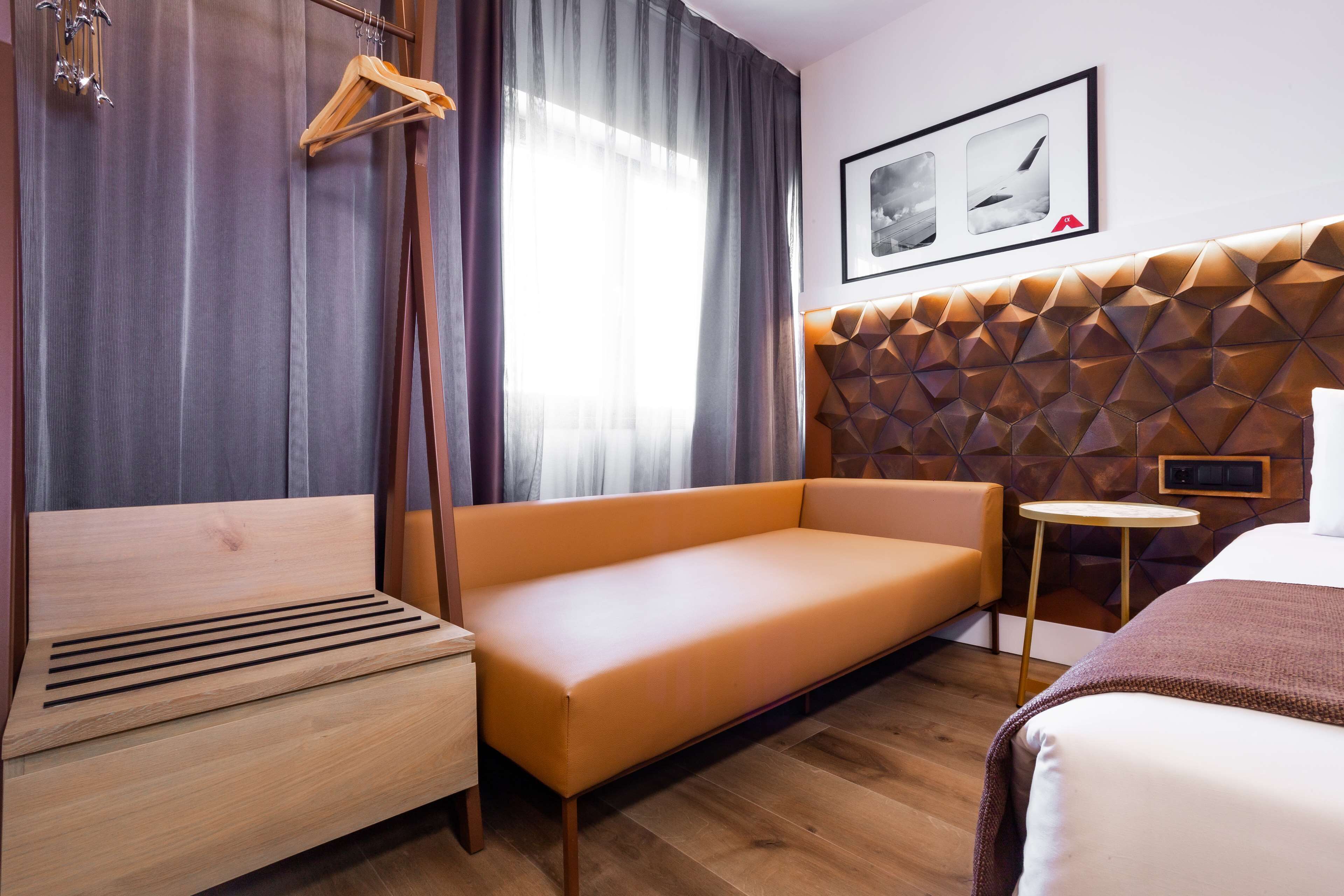 Una habitación de hotel moderna con un sofá de madera, una cama, una mesita redonda y un cabecero con estampado geométrico.