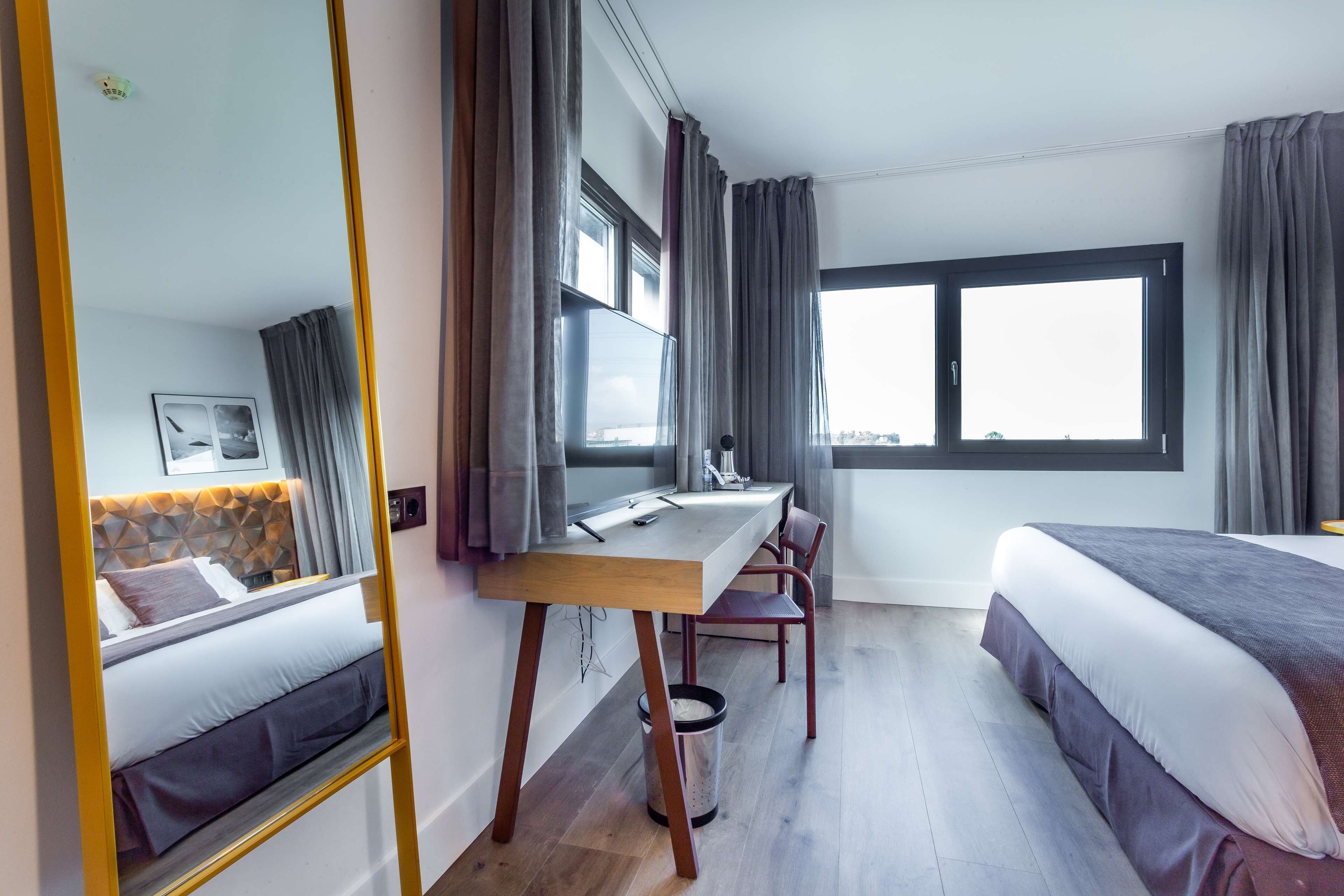Une chambre d’hôtel avec un lit, un bureau, une chaise, une grande fenêtre et un miroir.