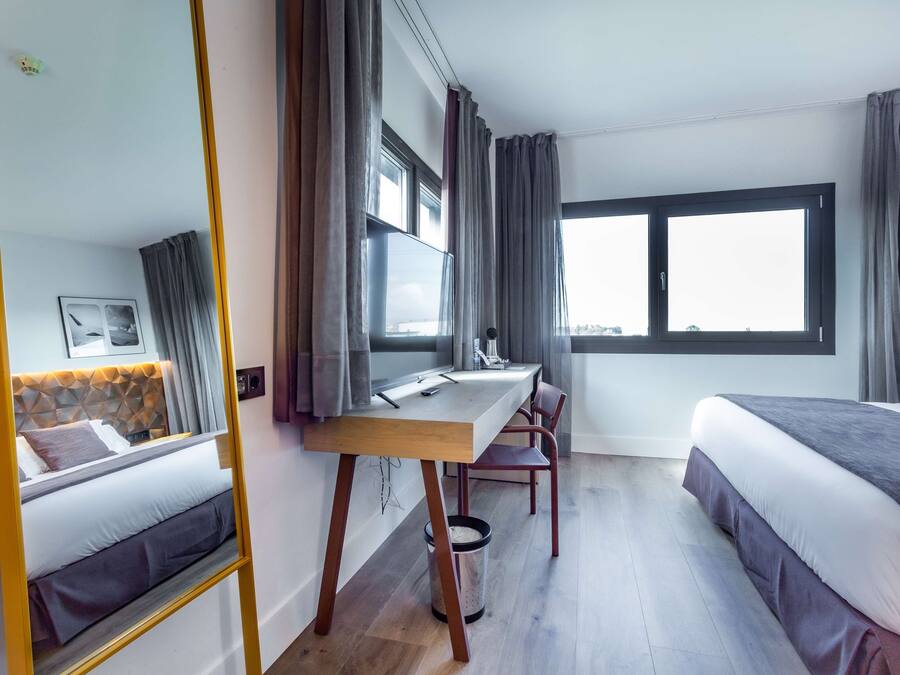 Une chambre d’hôtel avec un lit, un bureau, une chaise, une grande fenêtre et un miroir.