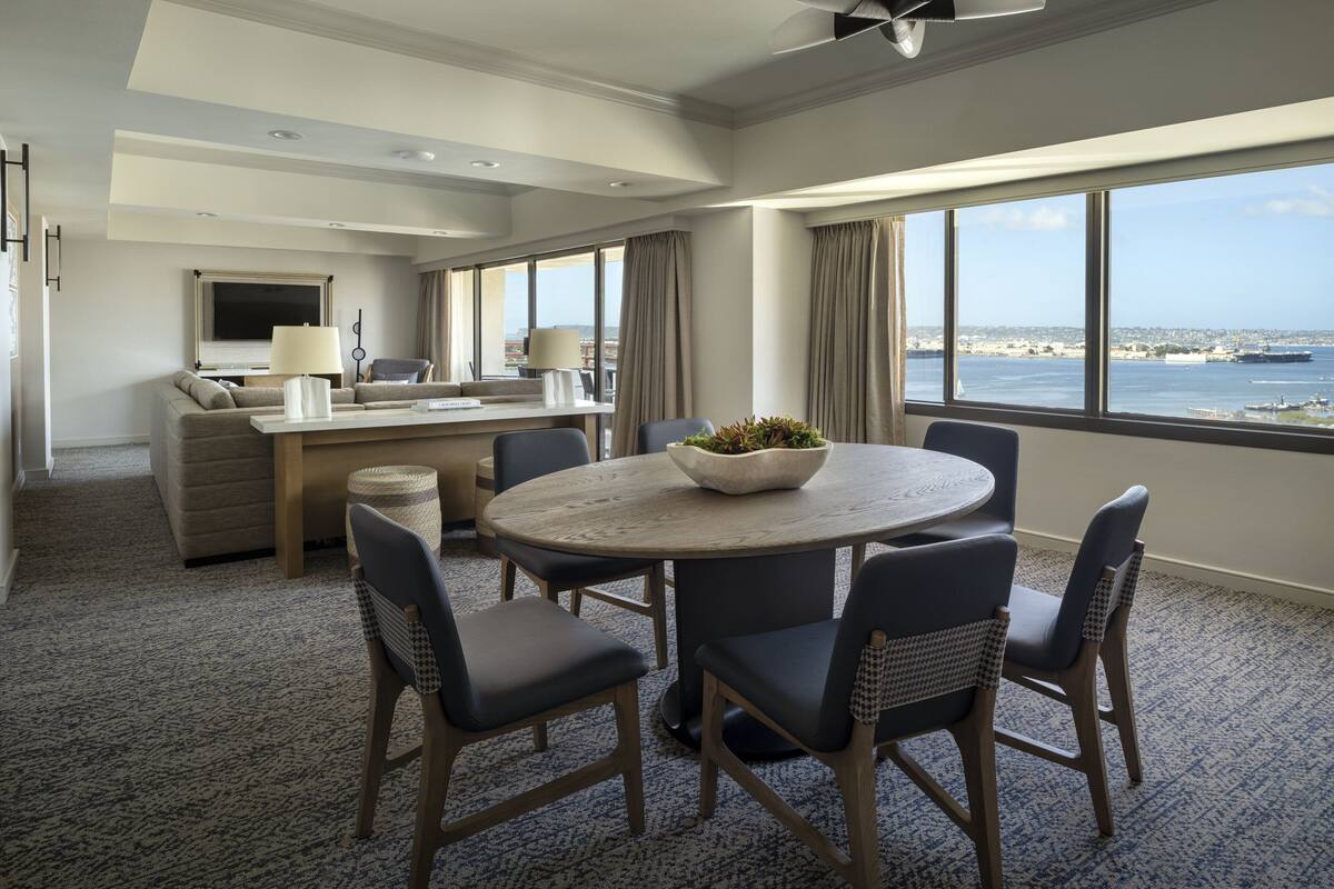 A modern hotel room with a dining area, a sofa, a TV, and a view of the sea.