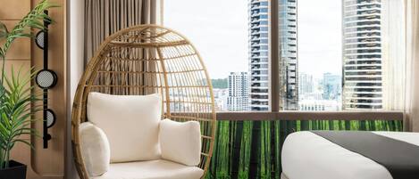 A hotel room with a large window, a wicker chair, a bed, and a potted plant.