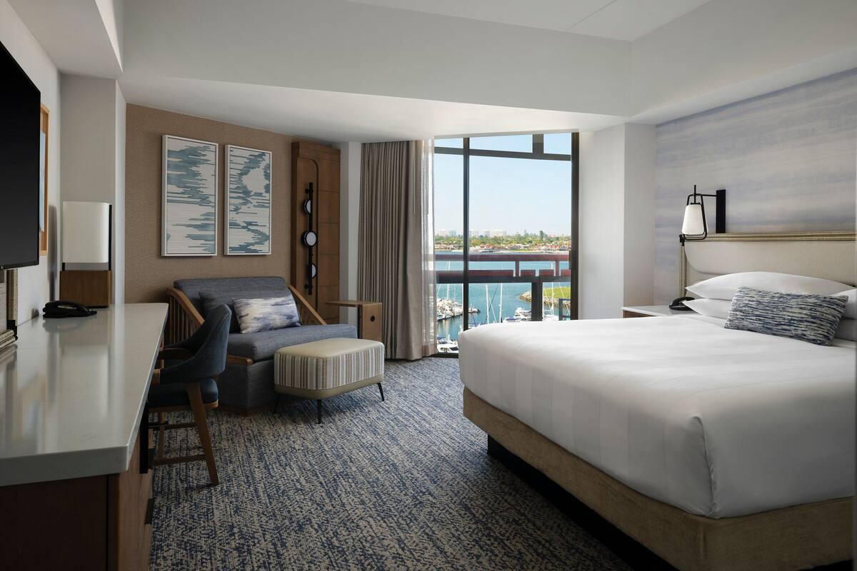 A hotel room with a bed, a desk, a chair, a TV, and a large window with a view of the water.