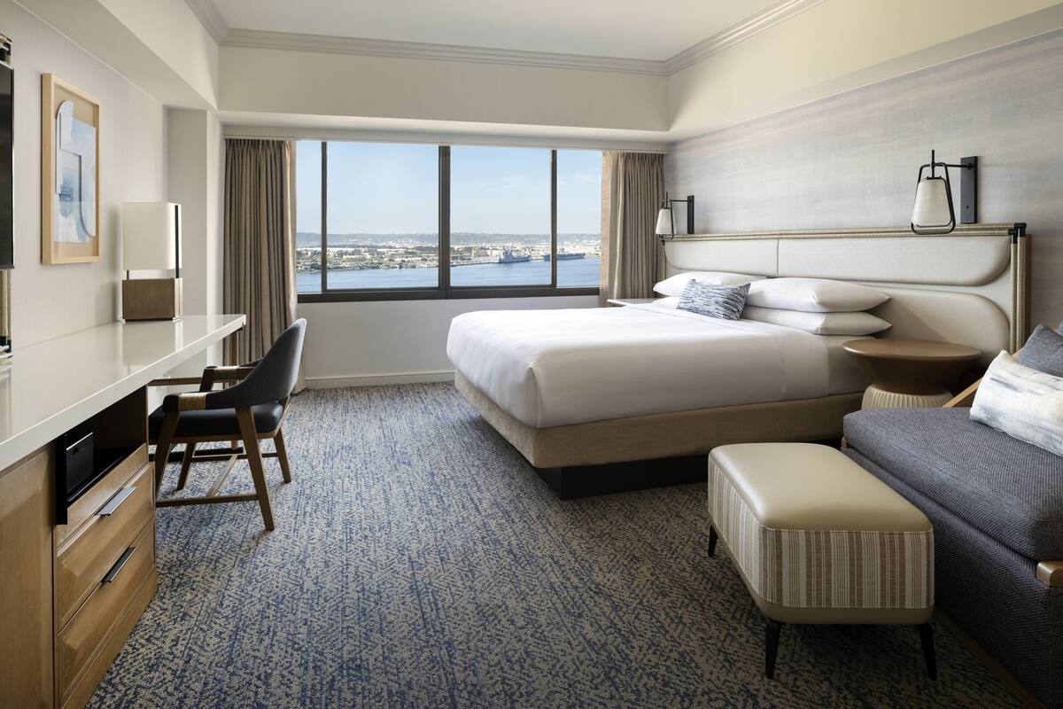 A hotel room with a large bed, a desk with a chair, a small ottoman, and a view of the city through a large window.